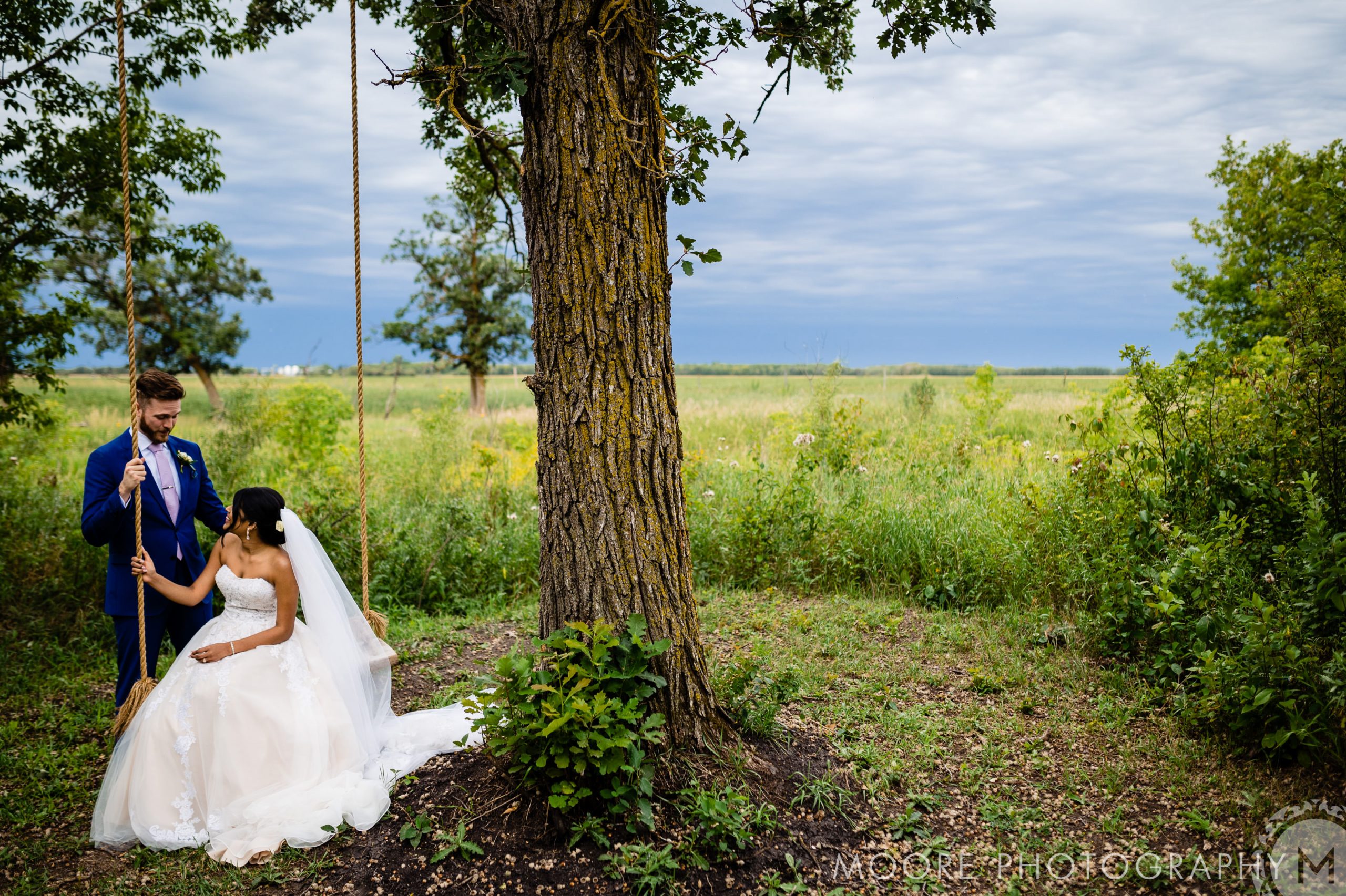 Bride on swing holds groom's hand in lush greenery, a scene fit for Winnipeg wedding venues.