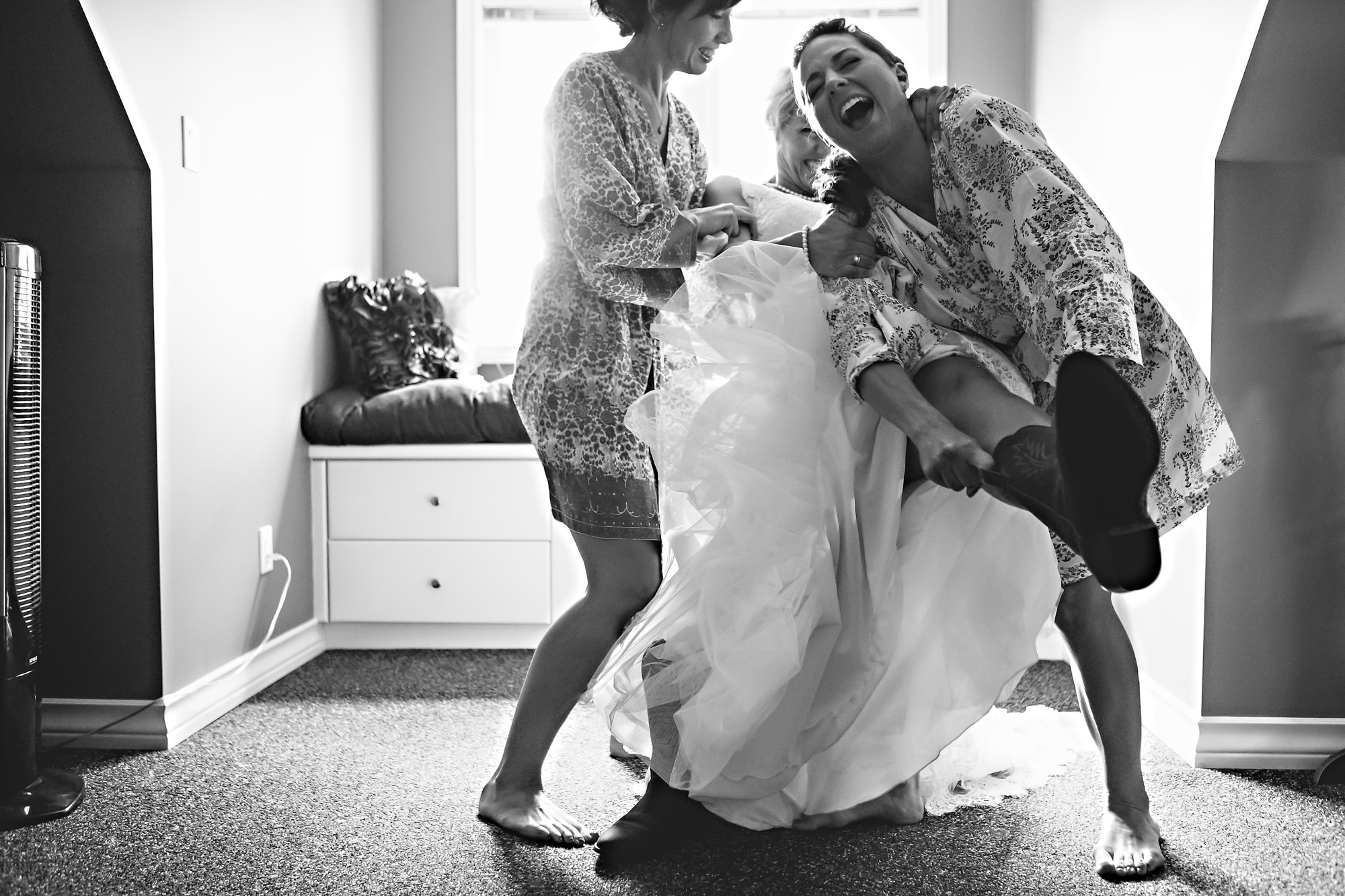 Three women in a room laugh and help each other. One is in a wedding dress, while the others assist in adjusting it. Captured by a Winnipeg Wedding Photographer, the scene is joyful and candid, with light streaming through a window in the background.