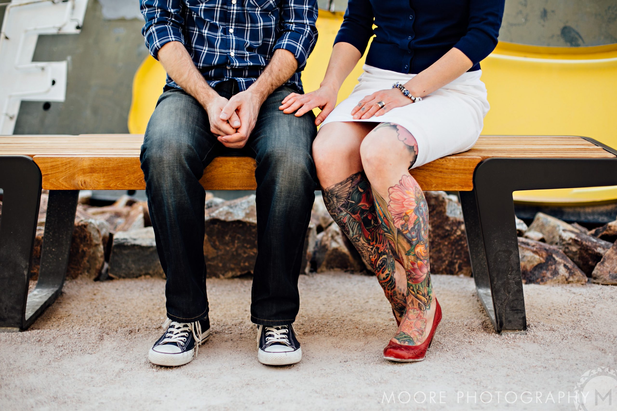 Two people in casual clothes on a bench, one's tattooed leg hints at destination wedding stories.