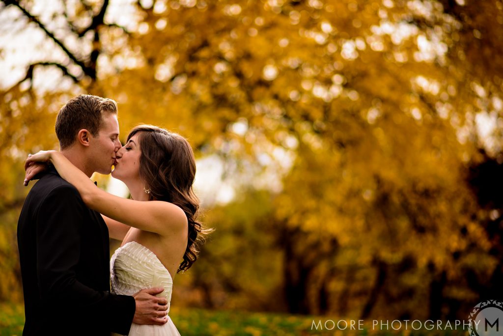 A bride and groom, embracing and kissing with fall leaves in the background