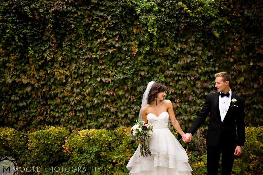 Bride and groom, holding hands and looking at each other in front of a green vine wall in Winnipeg Manitoba