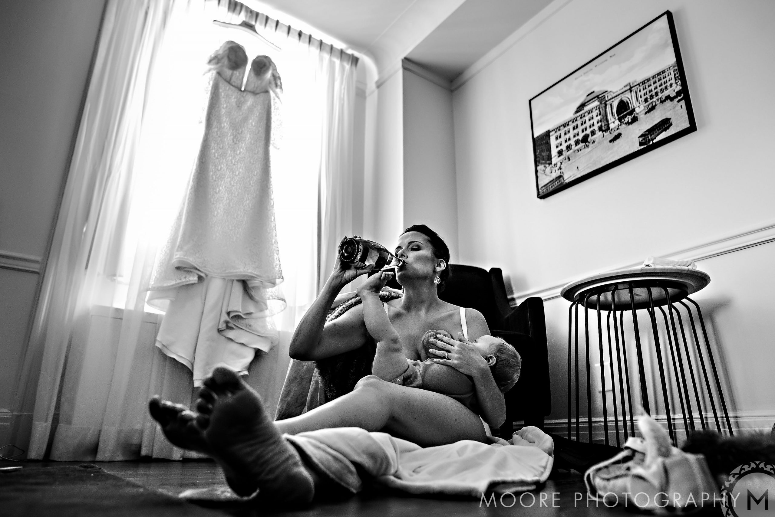 Bride in robe, sipping, holding baby; gown by window at Winnipeg wedding venue.