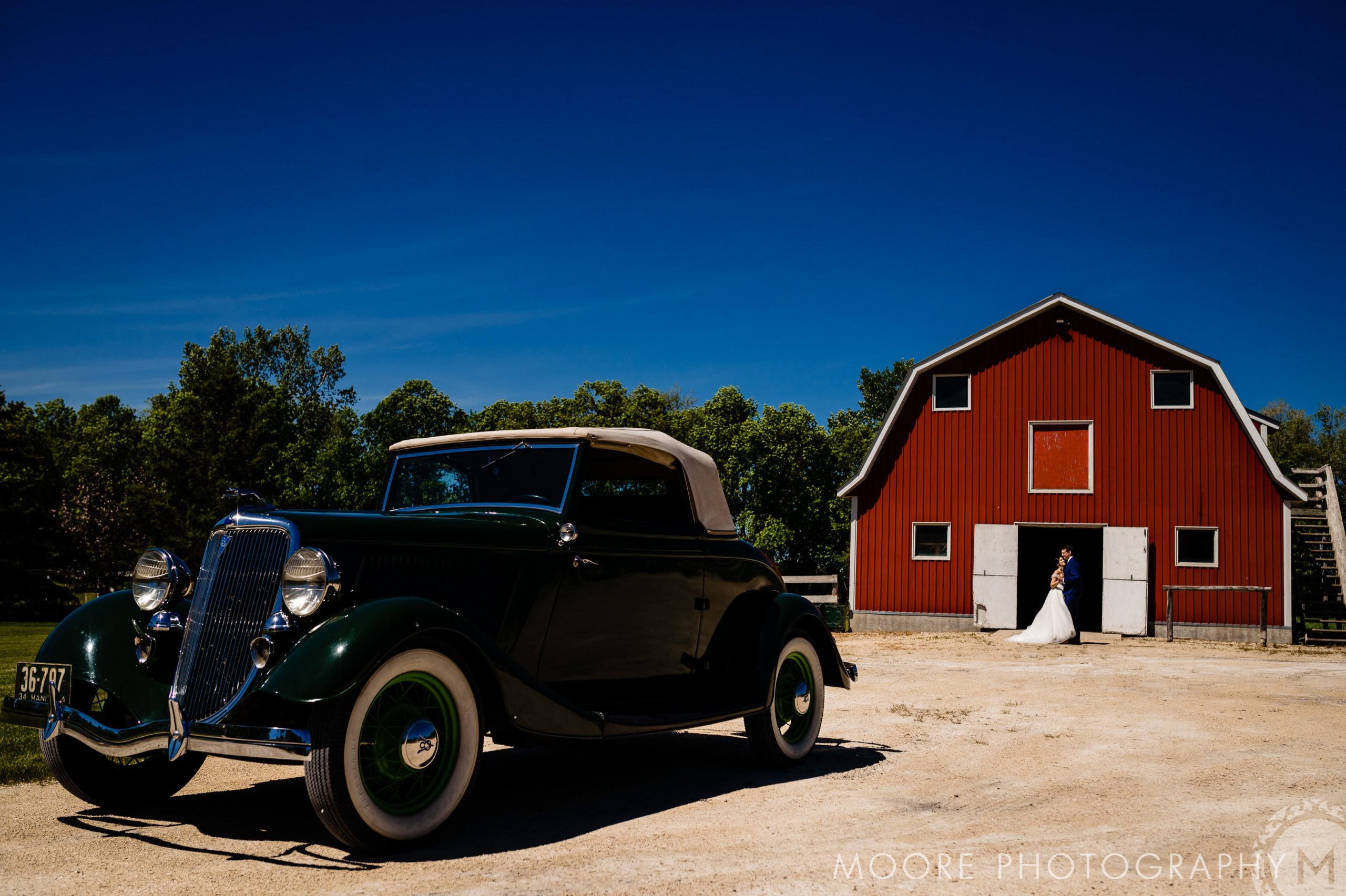 Vintage car by a red barn, bride awaits at Winnipeg wedding venues under clear blue sky.