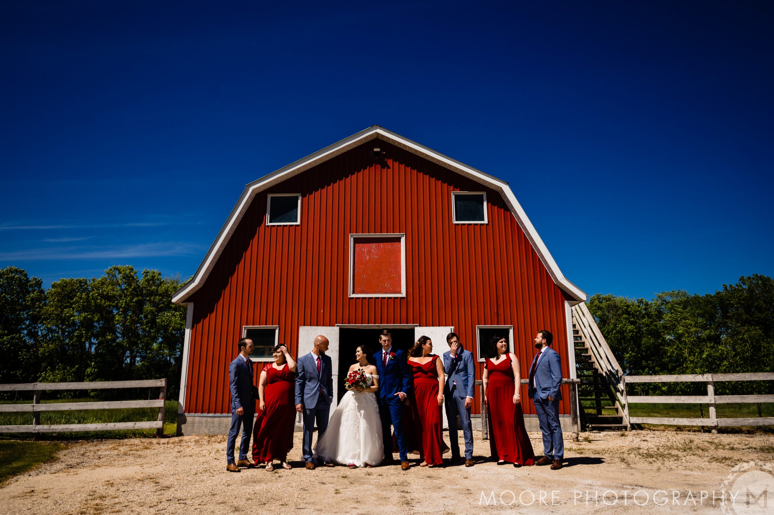 Wedding party in red and gray attire posing by a red barn on a sunny Winnipeg wedding day.