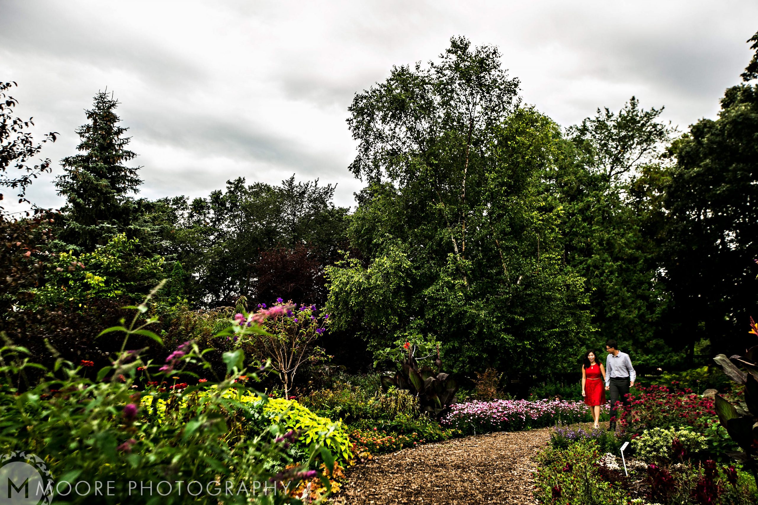 A relaxed couple strolls through a lush garden path under Winnipeg's cloudy sky.