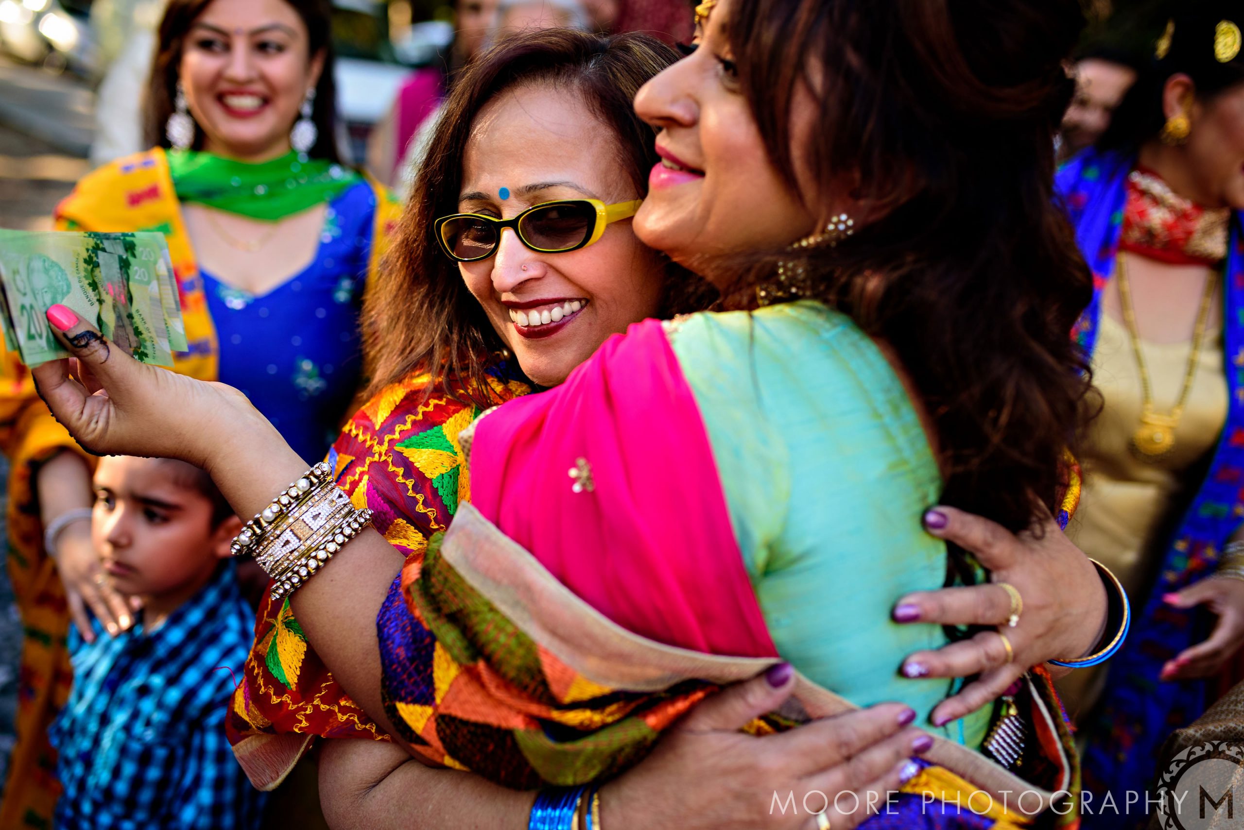 Women in colorful attire hugging at an Indian wedding in Winnipeg with onlookers nearby.
