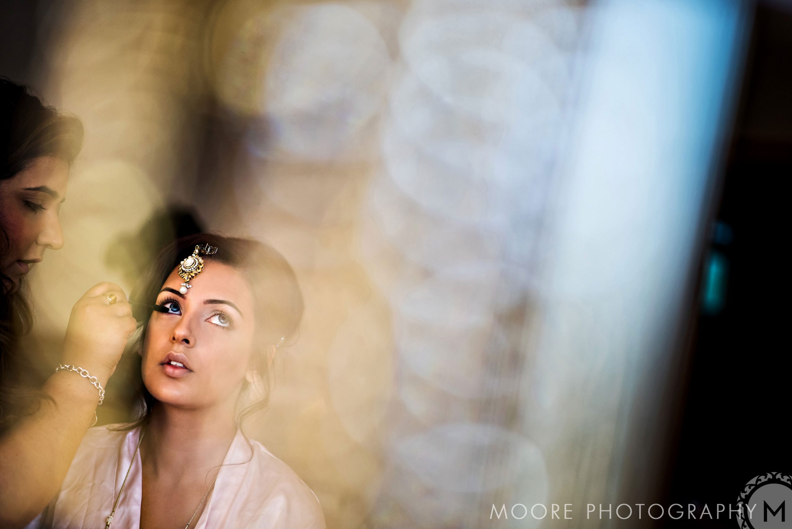 Woman getting makeup done, wearing a decorative headpiece, in soft Indian wedding lighting.