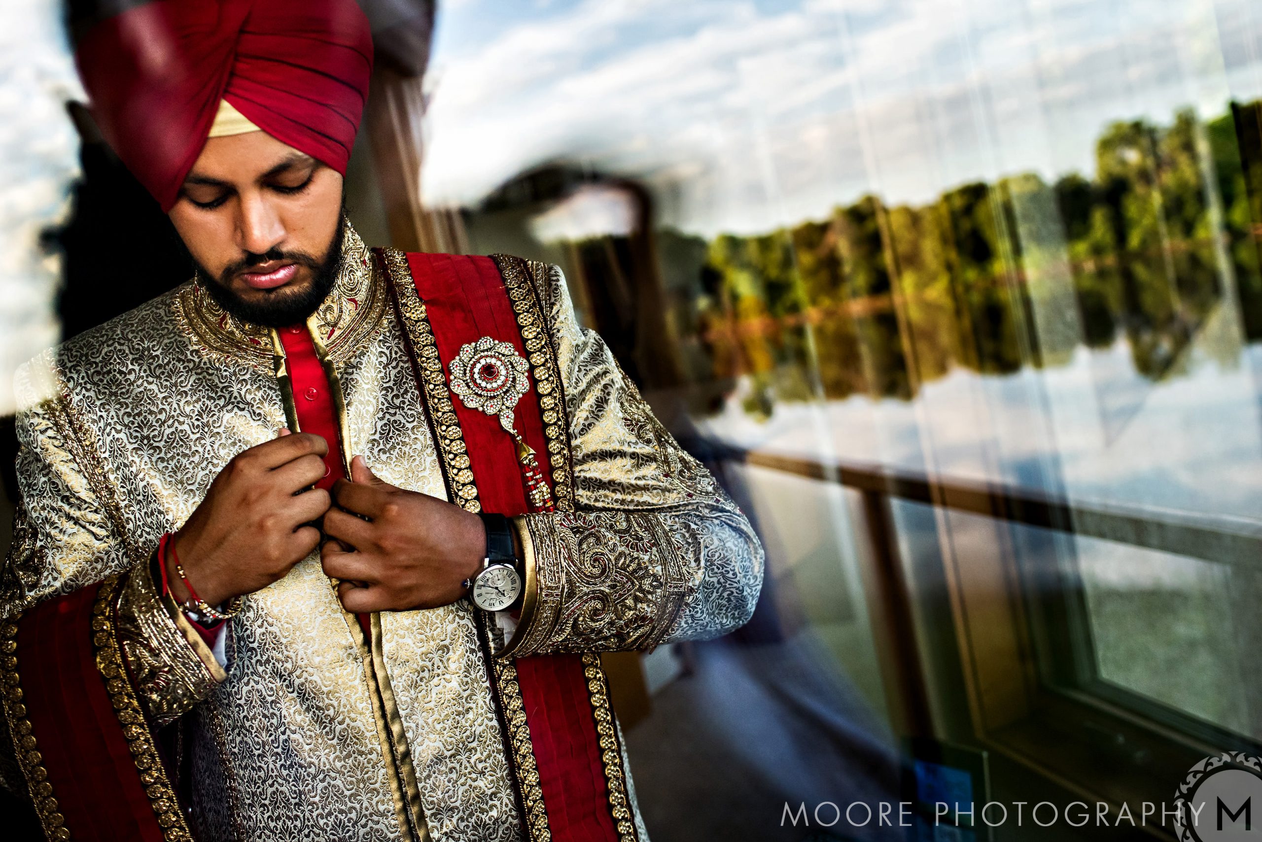 Man in ornate attire and turban, adjusting a button, reflected in a scenic Indian wedding view.