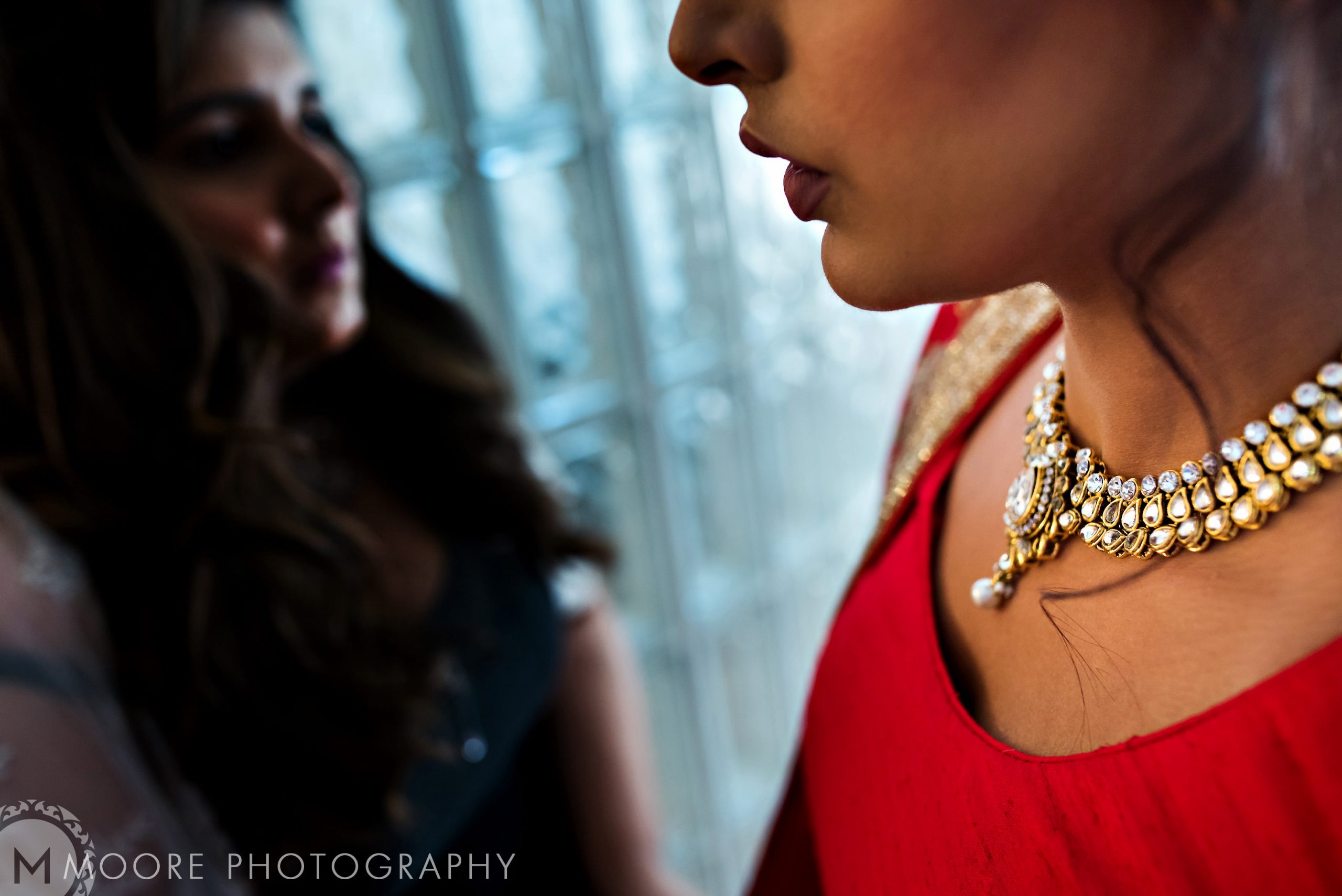 Close-up of a woman in red with pearls at an Indian wedding; another woman is blurred behind.