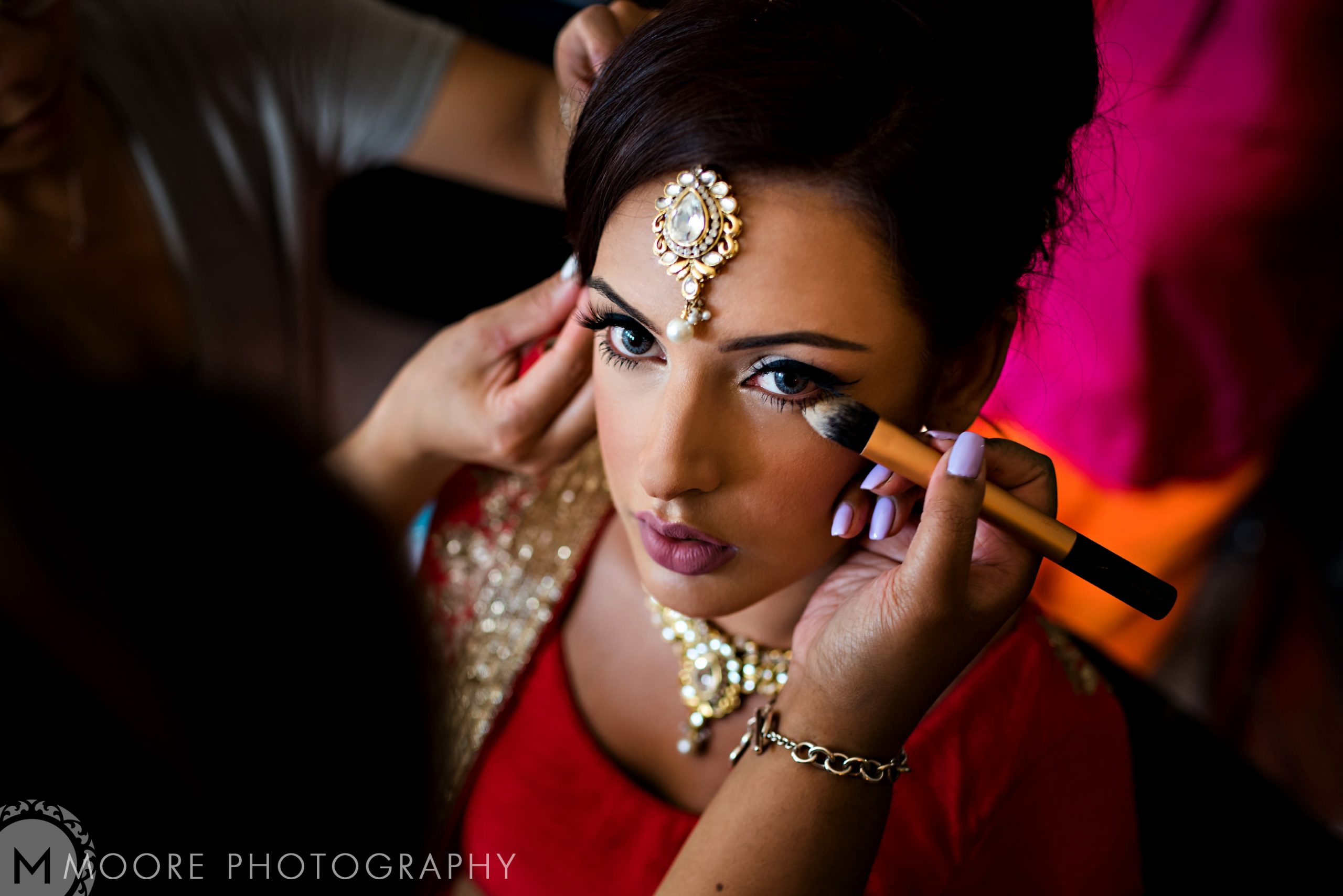 Woman getting makeup applied, wearing traditional jewelry and a red Indian wedding outfit.