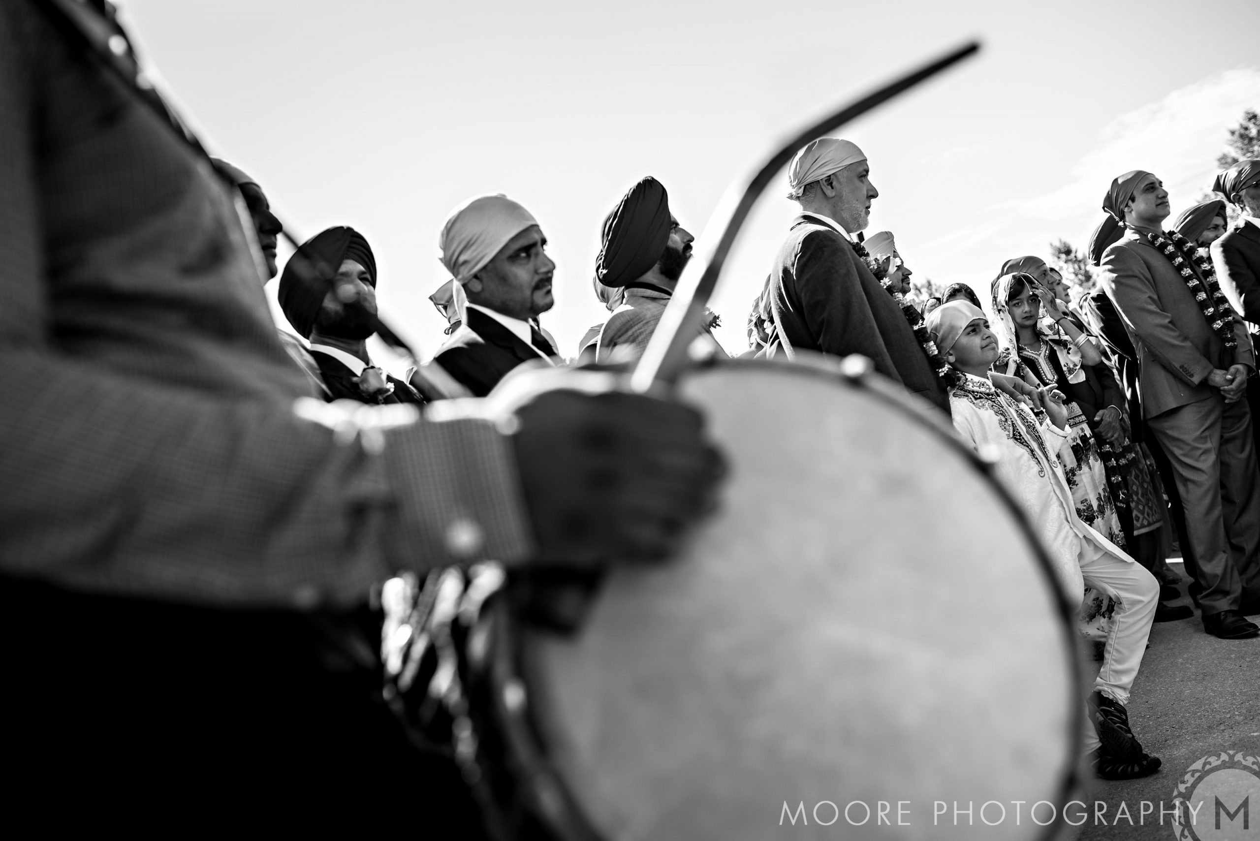 Black and white image of men in turbans and suits at an Indian wedding, one playing a large drum.