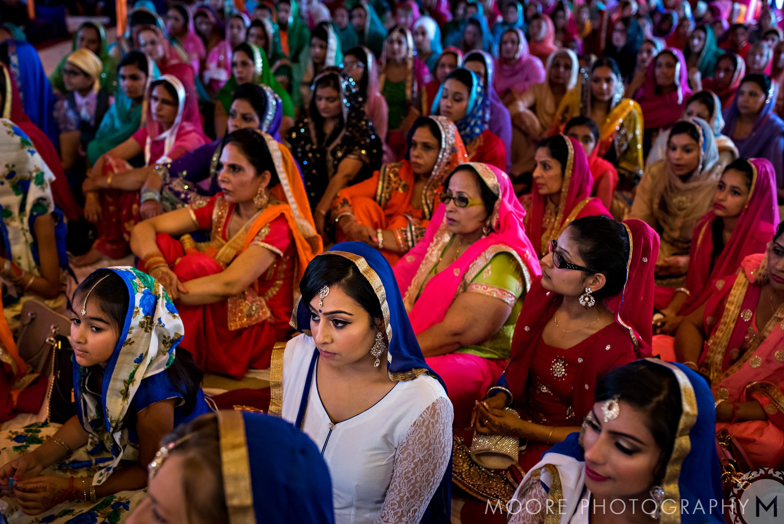 A large group of women and children sit together wearing colorful, traditional Indian wedding attire.