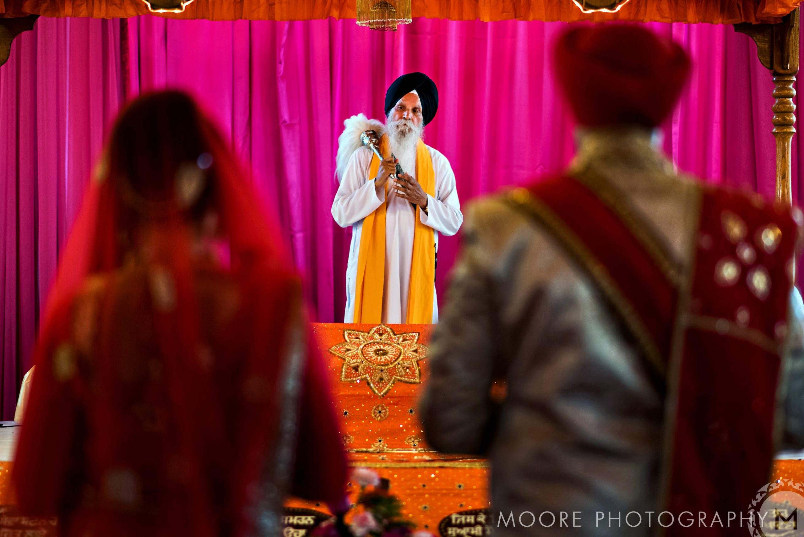 Indian wedding ceremony with a priest, vibrant pink and orange drapes in the background.