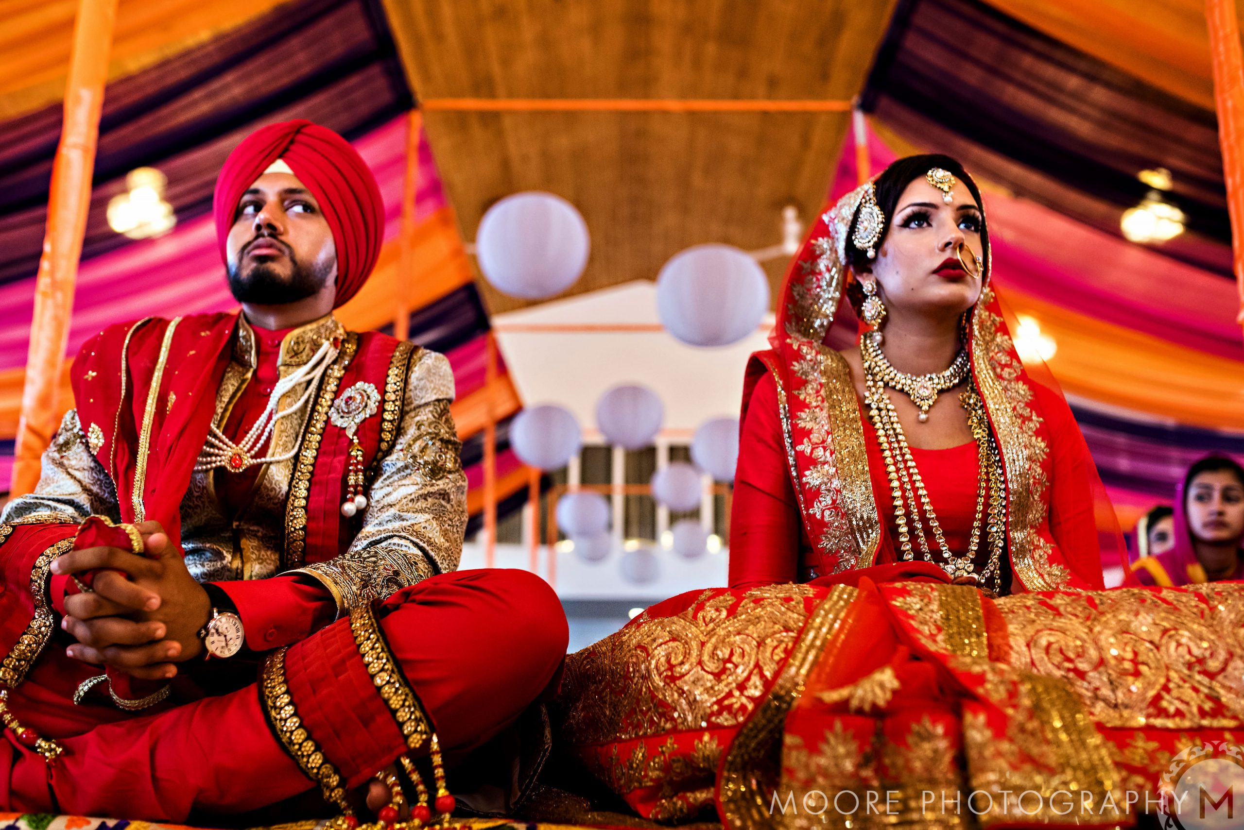 An Indian wedding couple sits indoors under colorful drapes in traditional Sikh attire.
