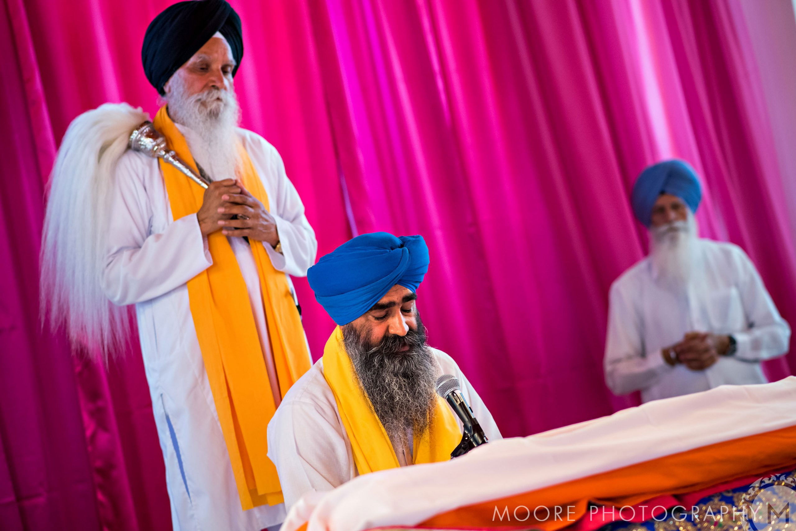A man in a blue turban sits by a microphone, with two others behind at an Indian wedding.