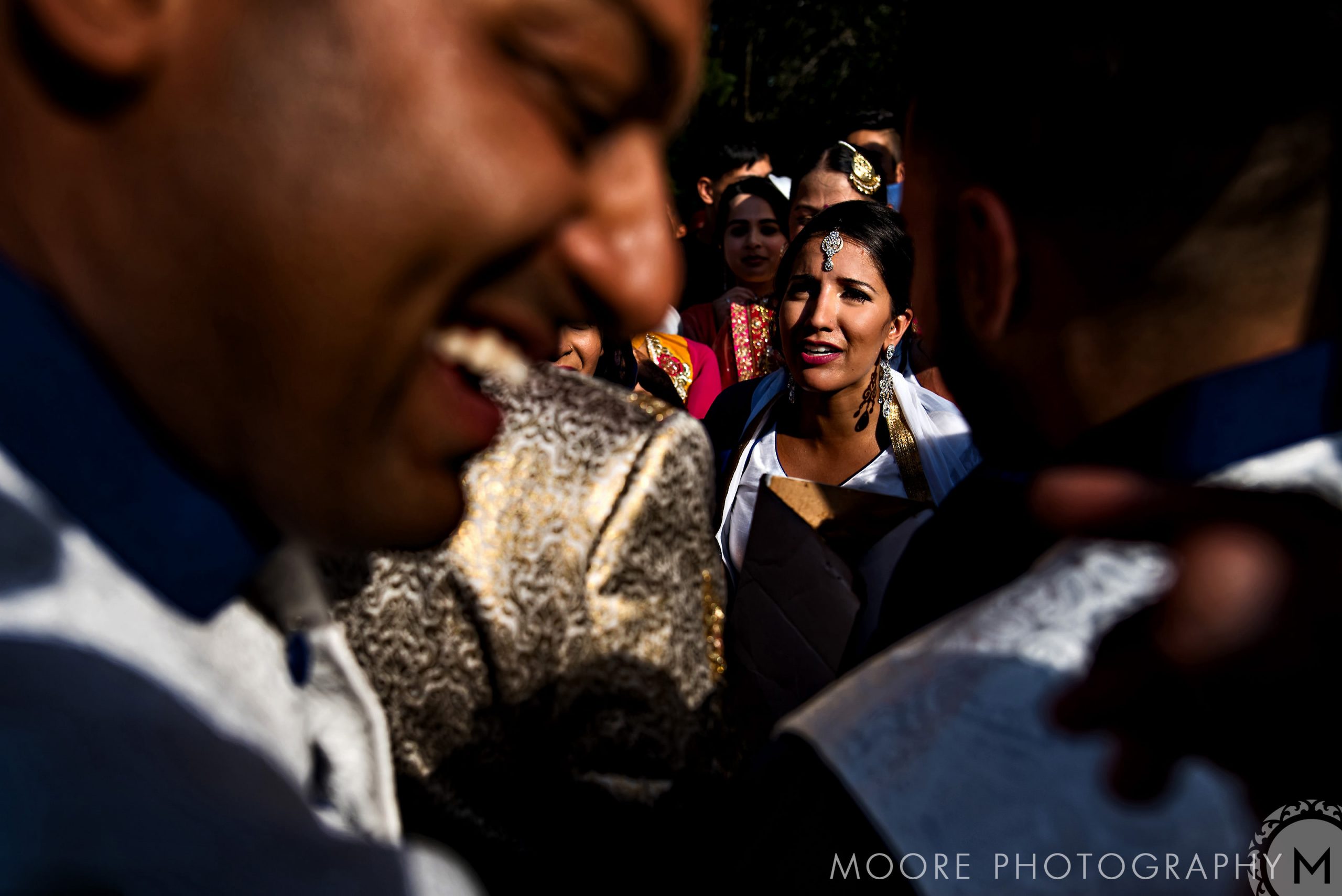 A woman in bright attire smiles as people celebrate an Indian wedding outdoors.
