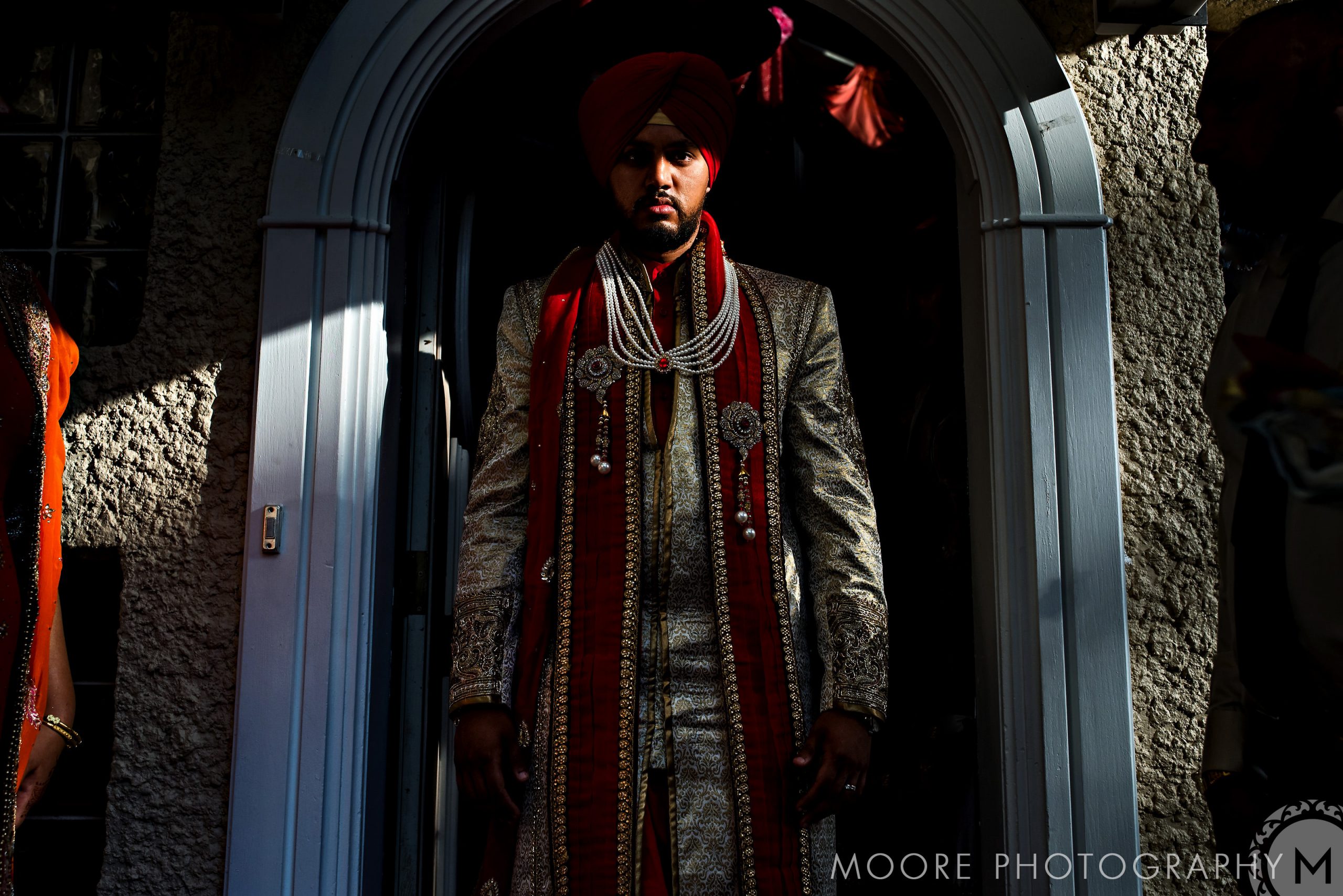 Man in traditional Indian wedding attire and red turban stands in an arched doorway, dramatic lighting.