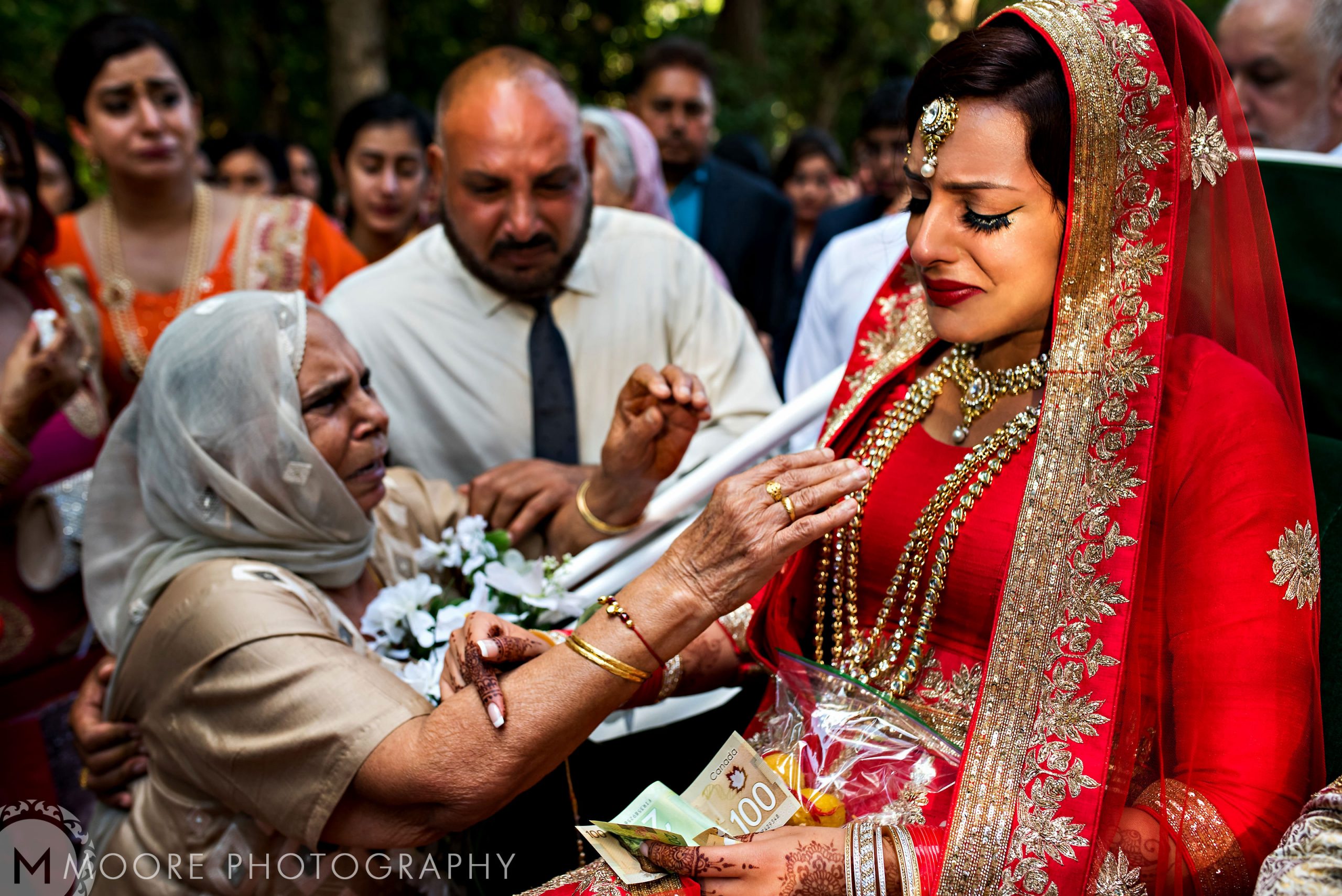 Bride in red crying, hugged by elderly woman in traditional Indian wedding attire.