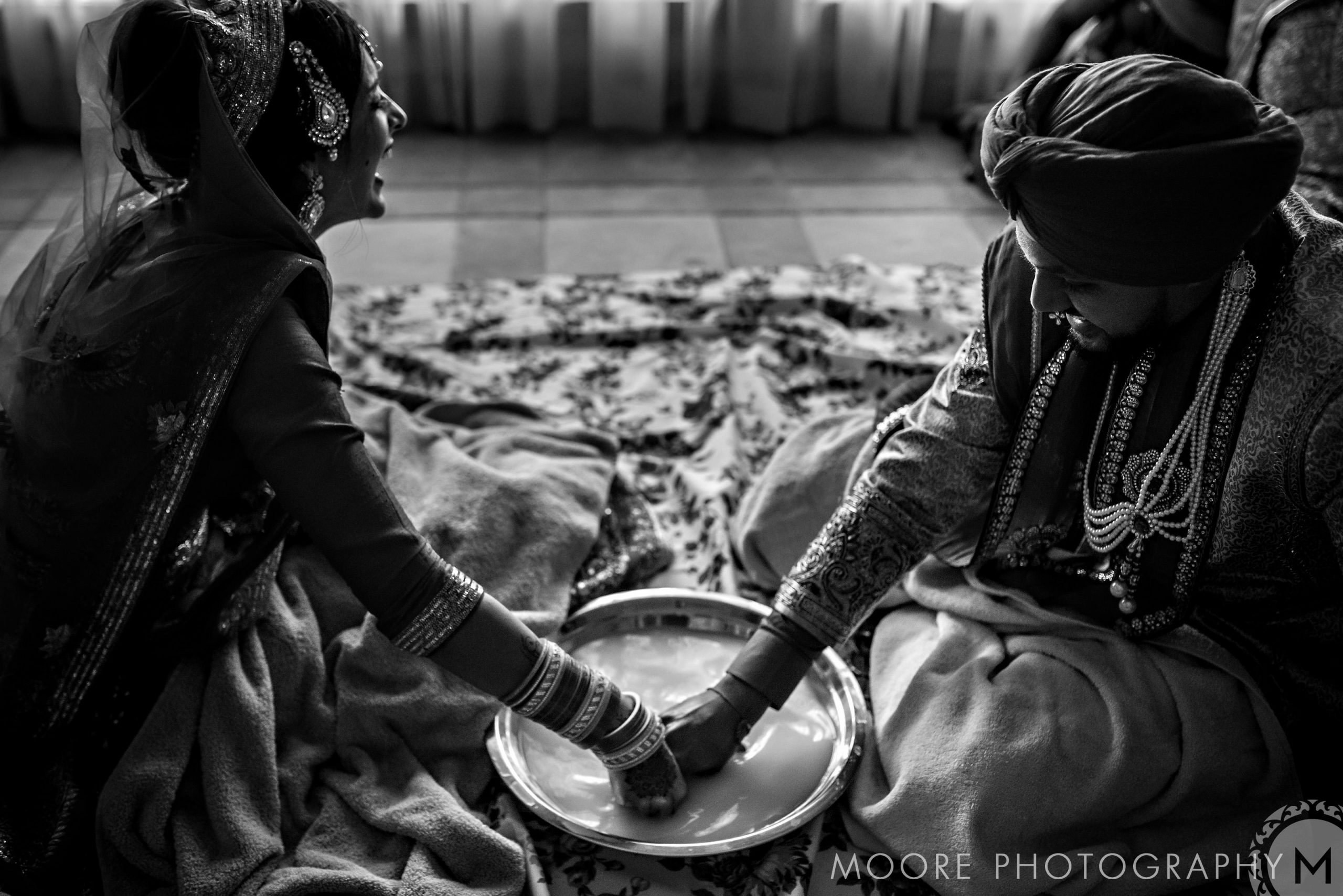 A couple joins an Indian wedding ritual, hands immersed in a bowl of water.