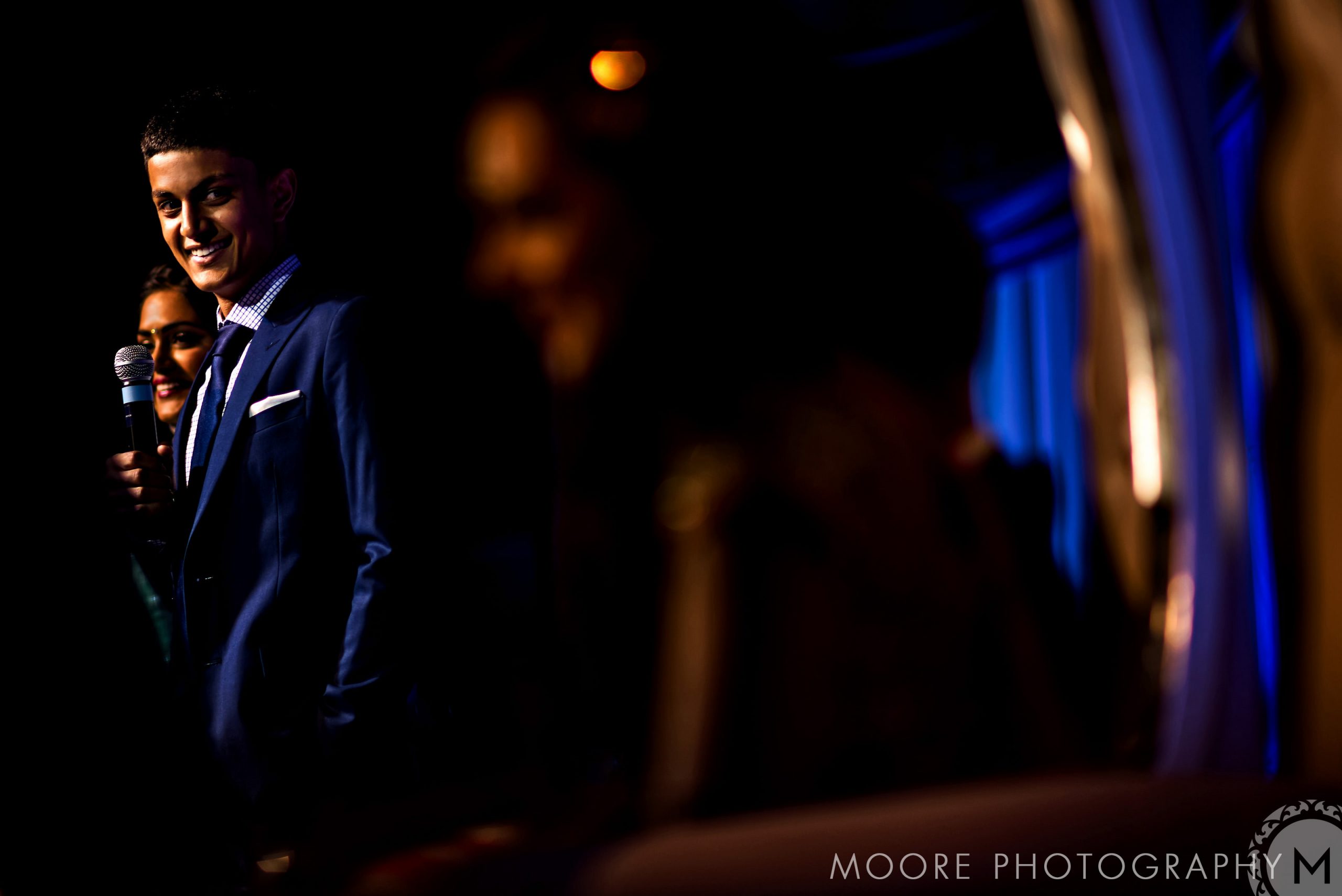 Man in a blue suit smiles, holding a microphone at an Indian wedding on a dimly lit stage.