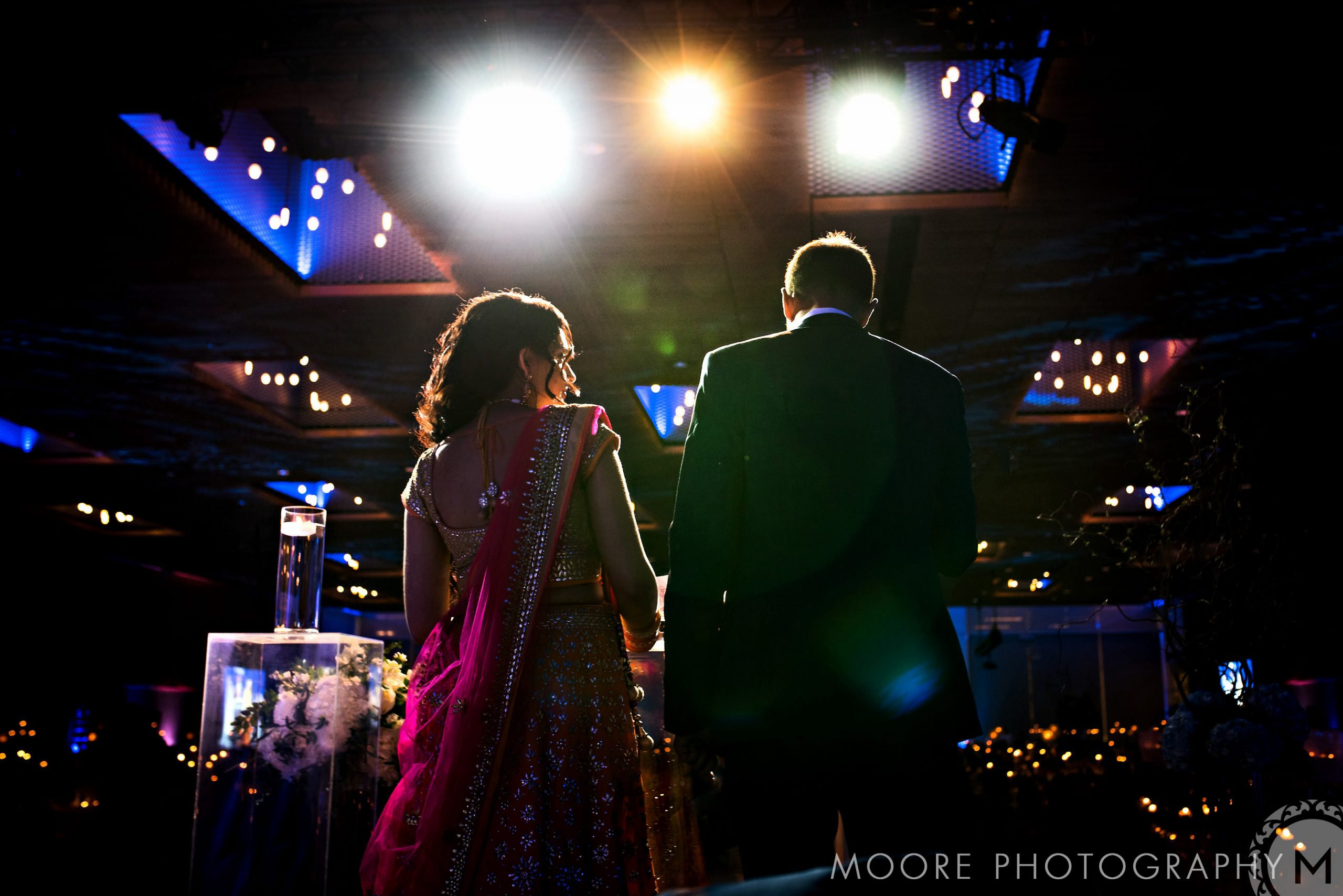 A couple in formal attire stands under bright lights at a glamorous Indian wedding event.