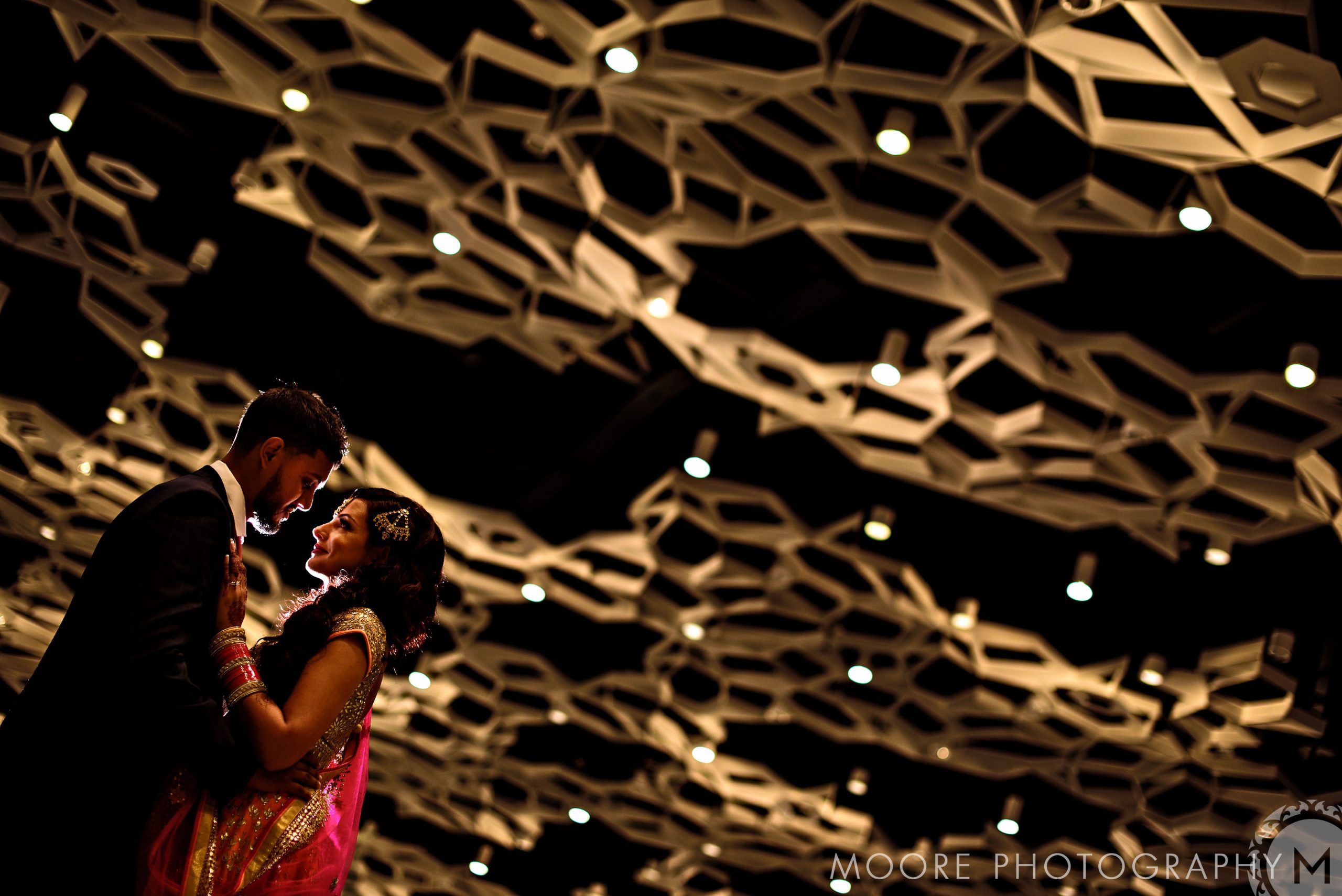 Couple embracing under a warm-lit, geometric ceiling at an Indian wedding.