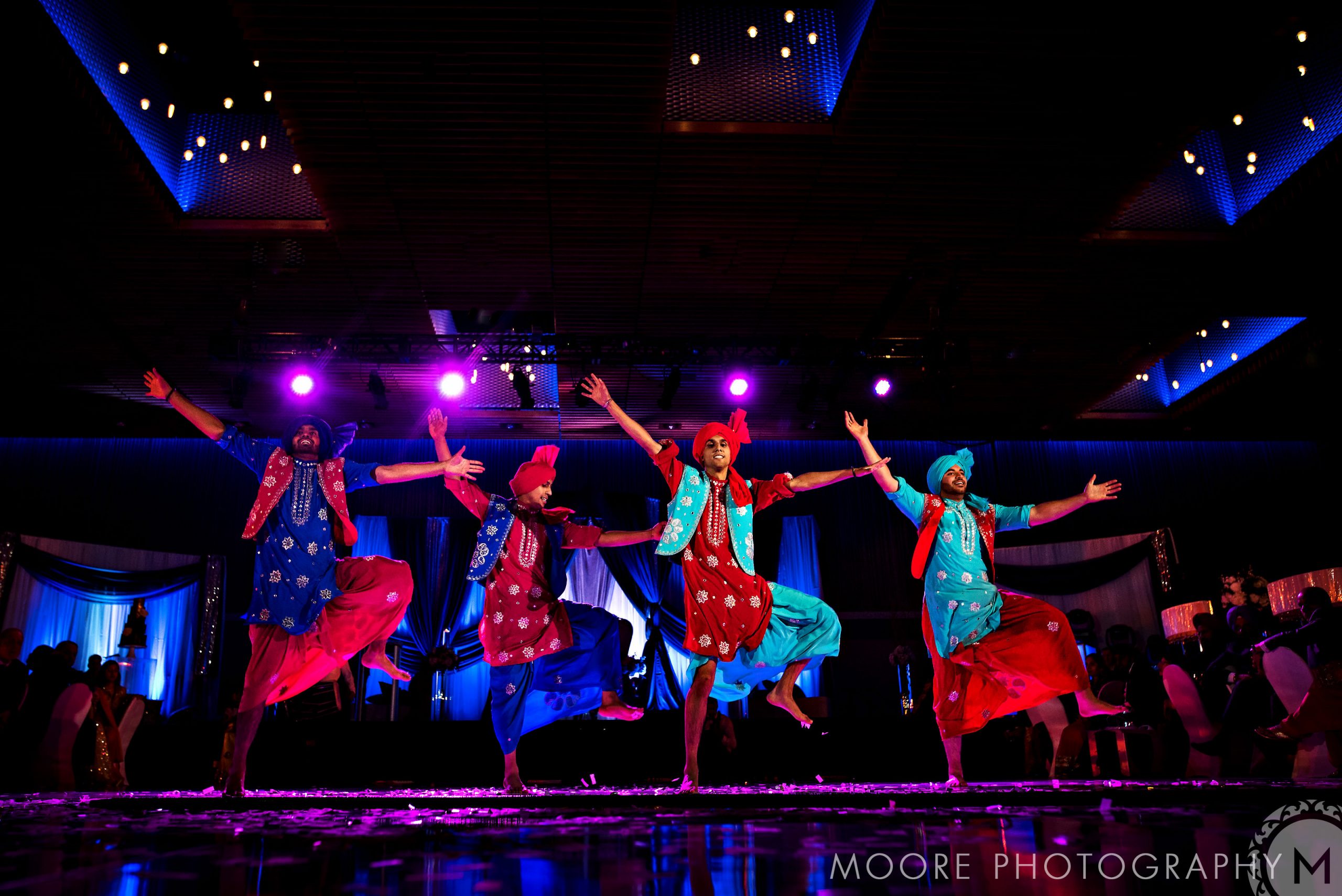 Dancers in vibrant costumes perform energetically at an Indian wedding stage.