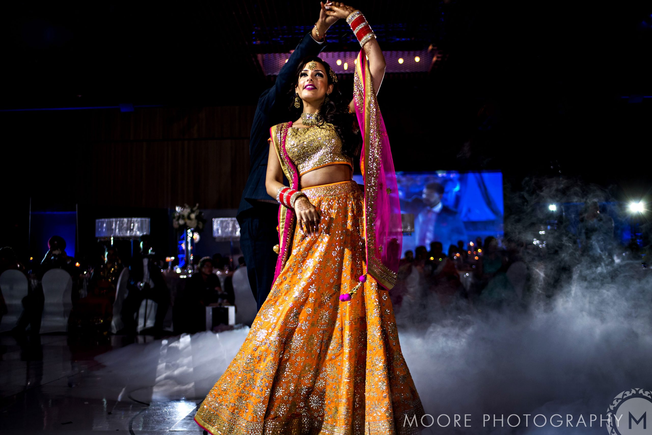 A person dances joyfully in a vibrant, traditional outfit at an Indian wedding.