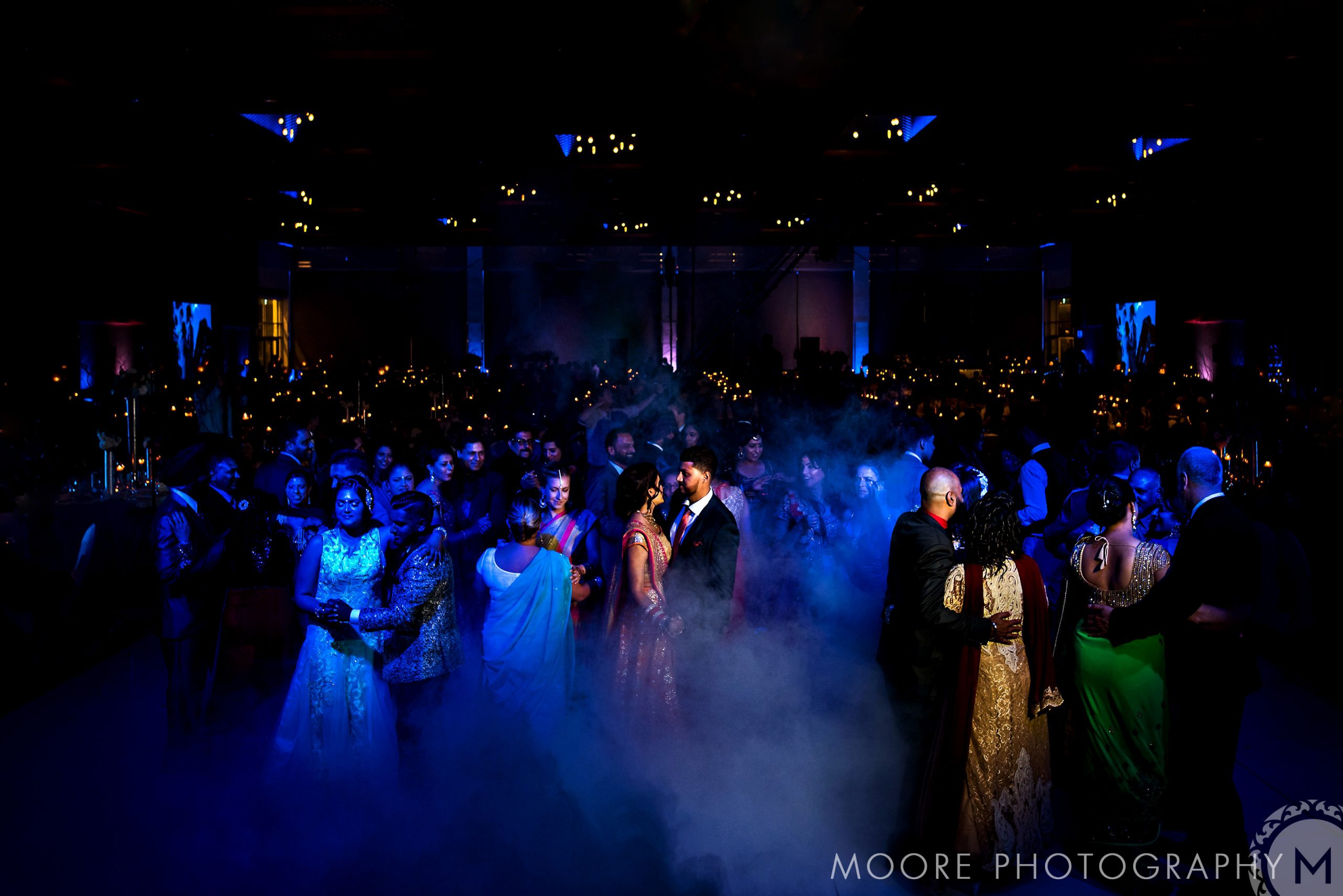 A crowded dance floor with colorful lights and fog at an Indian wedding.
