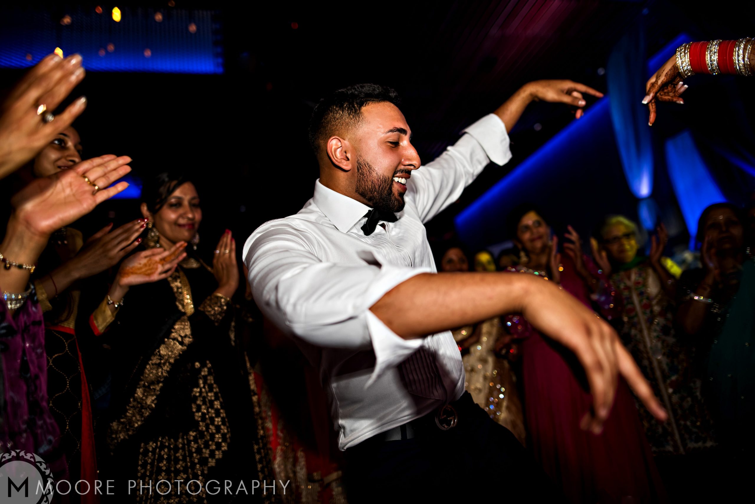 Man joyfully dancing at an Indian wedding, with others clapping around him.