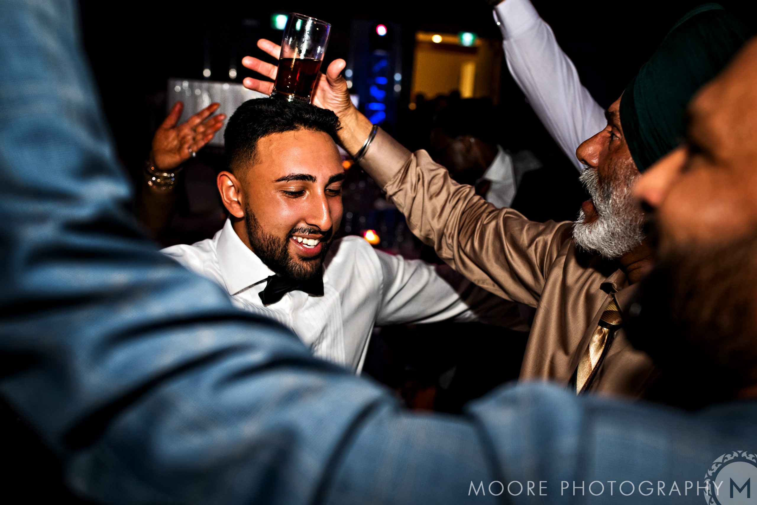 At an Indian wedding, people dance energetically, drinks in hand, looking joyful and festive.