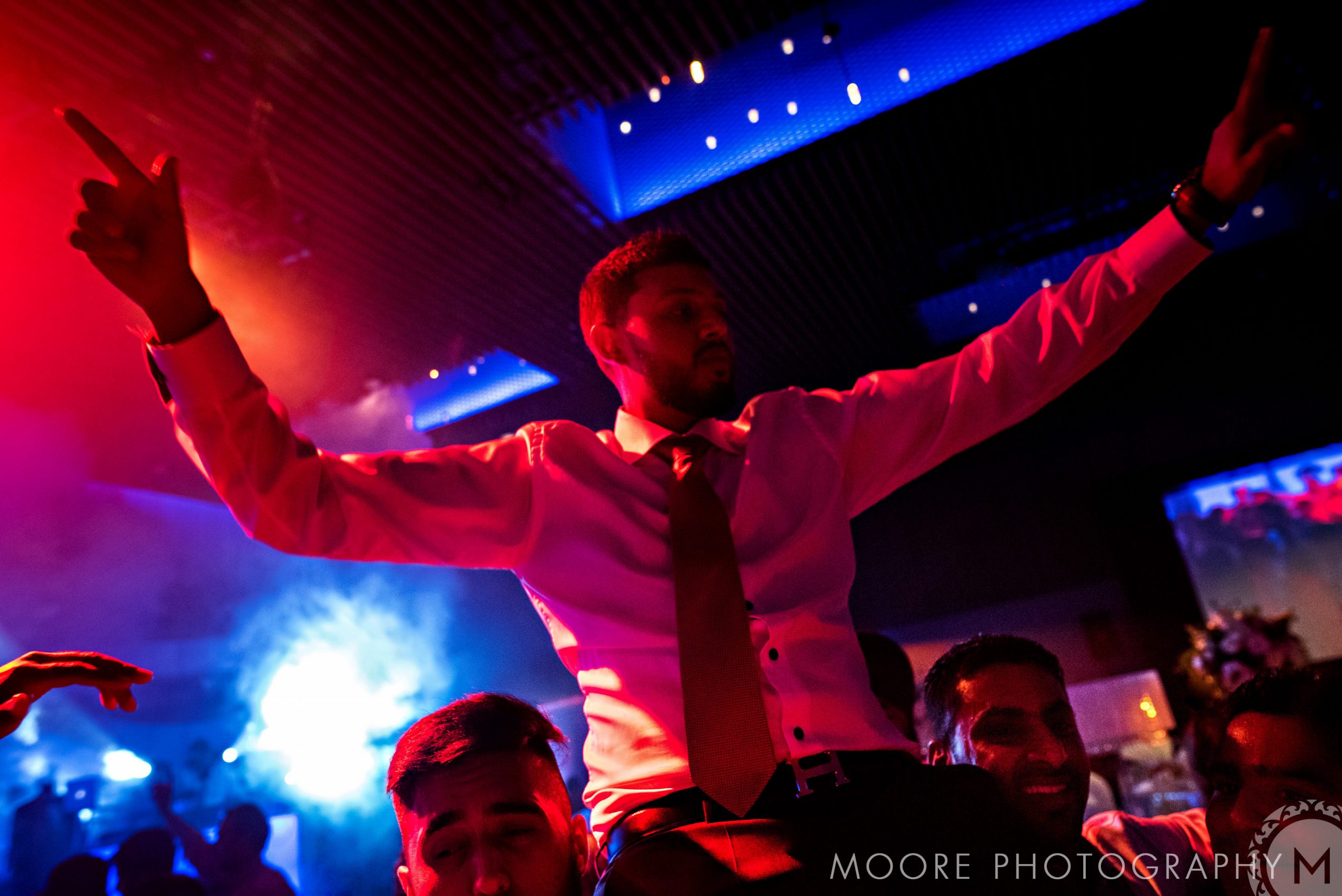 Man in white shirt and red tie being lifted at a lively Indian wedding under colorful lights.