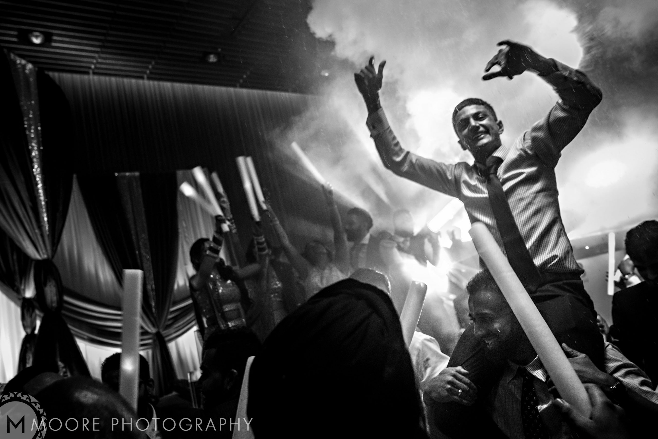Man celebrating on shoulders at a lively Indian wedding with lights and fog.