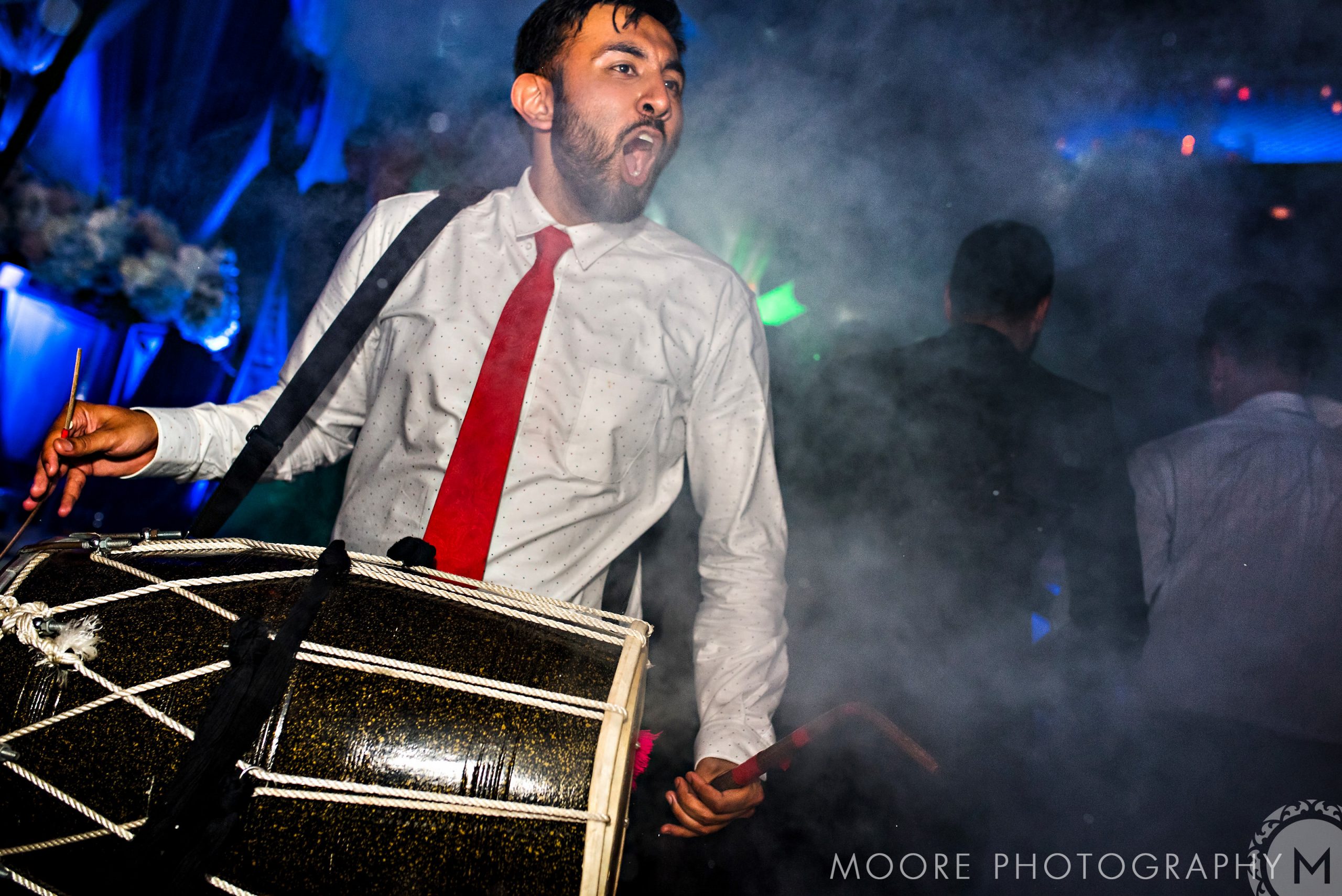 Man energetically playing a drum at an Indian wedding amid smoky atmosphere and blue lighting.