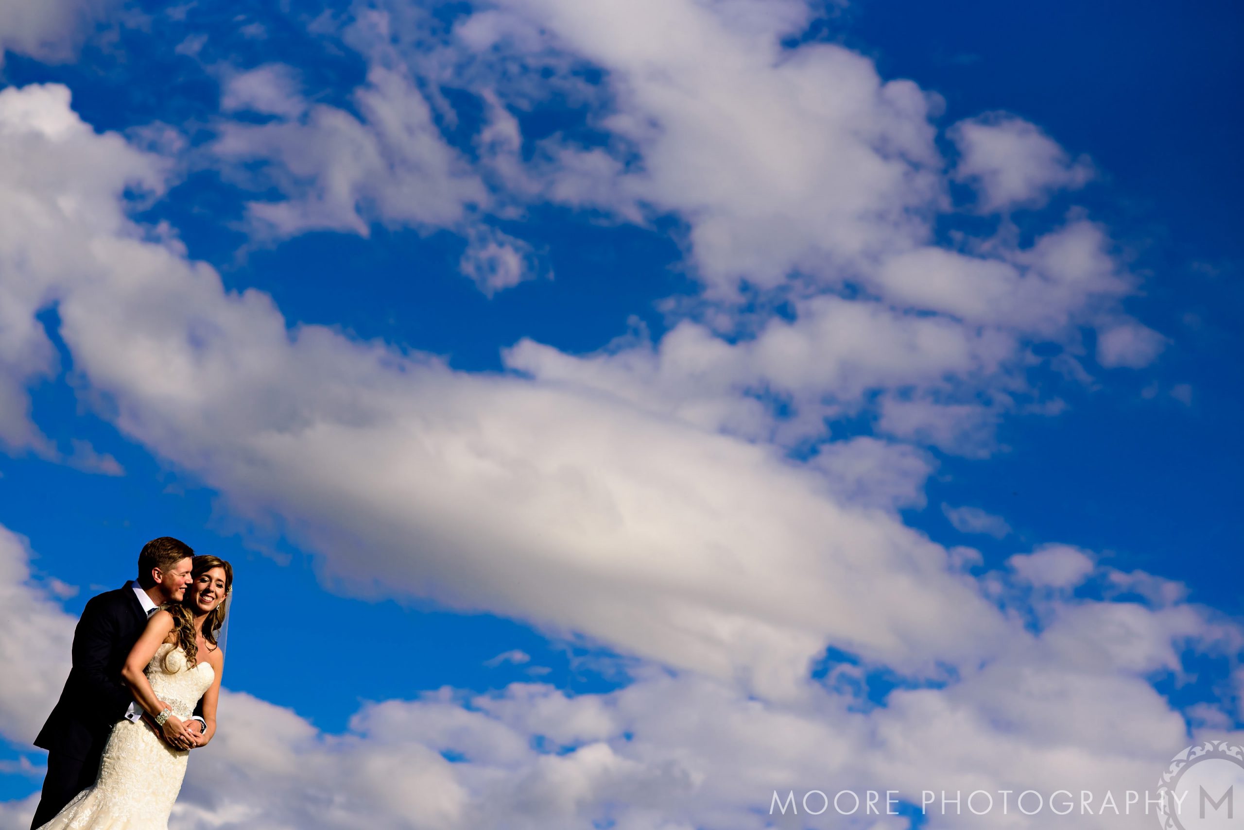 Bride and groom embracing under Winnipeg's bright blue sky with scattered white clouds.
