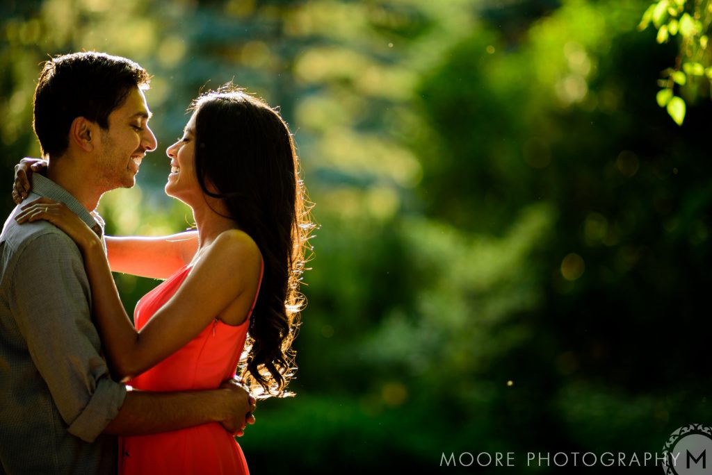 And engaged couple, embracing, looking into each other, eyes, and smiling with trees in the background during sunset at Assiniboine Park