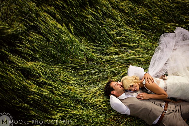 A couple lies on a grassy field in this serene image captured by a Winnipeg wedding photographer. The woman, in a white gown, rests her head on the suited man's chest as the grass swirls around them, forming a heart shape. The scene exudes peace and intimacy.