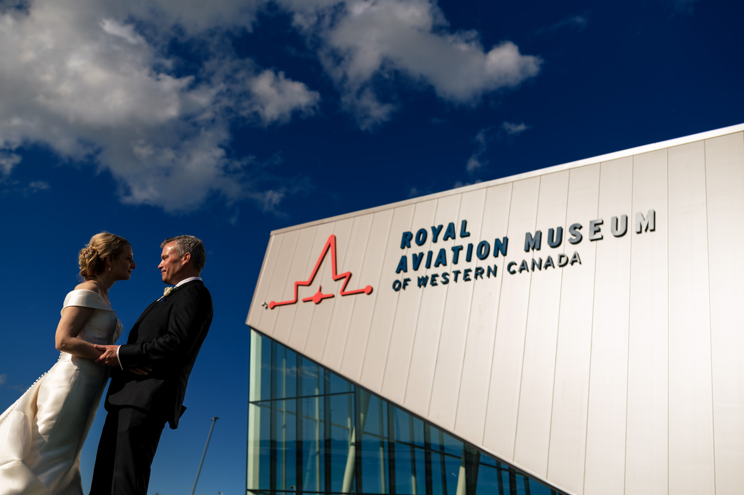 Couple embracing outside Winnipeg wedding venue under a blue sky at the Aviation Museum.