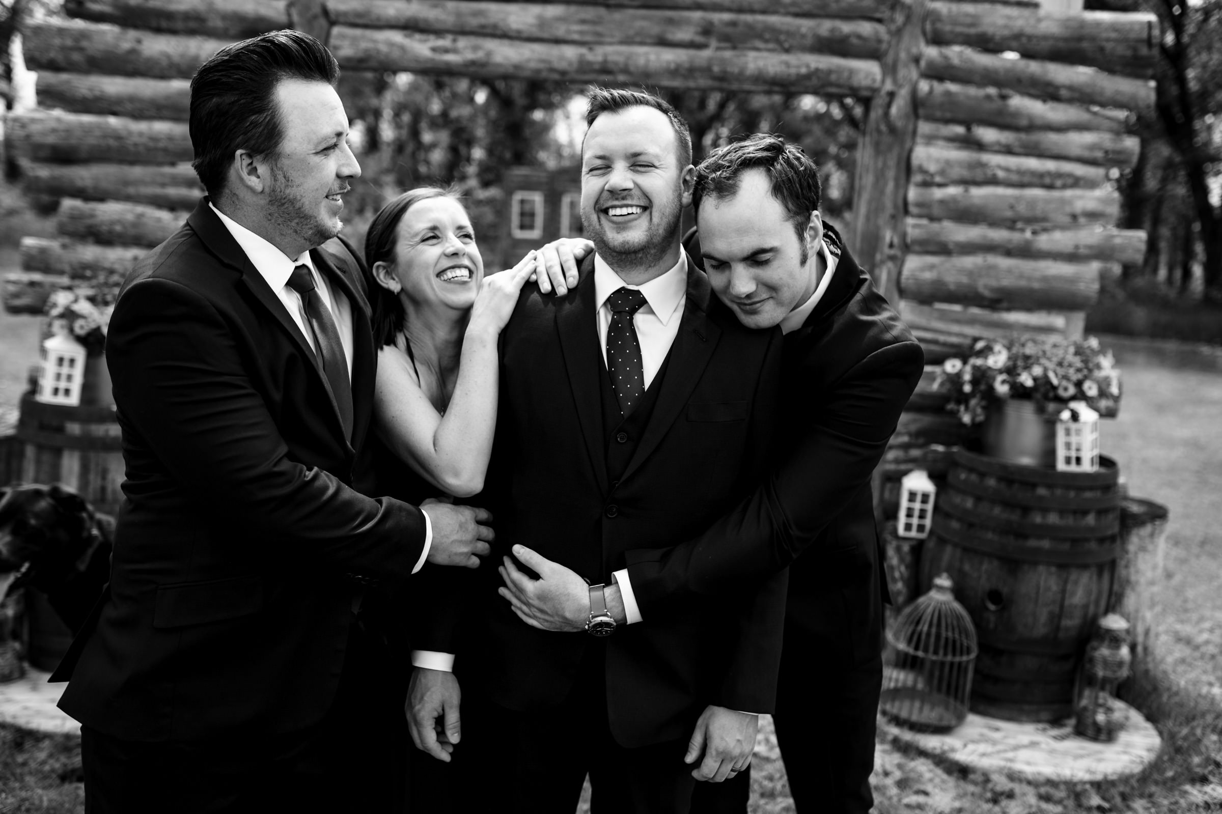 Four people in formal wear laugh and hug near wooden barrels at Winnipeg wedding venues.