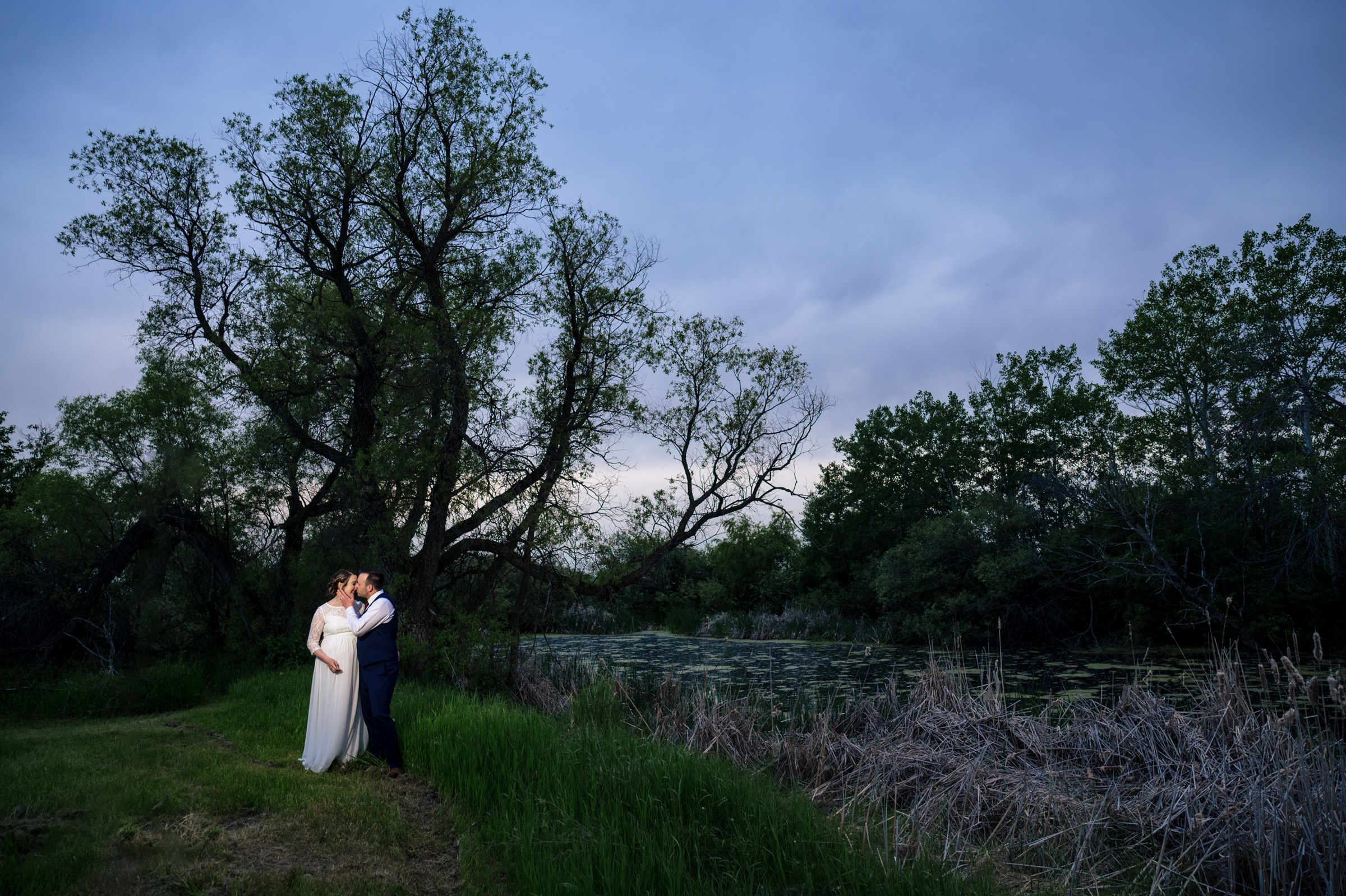 Couple embracing by a pond and trees at twilight, a serene Winnipeg wedding venue.