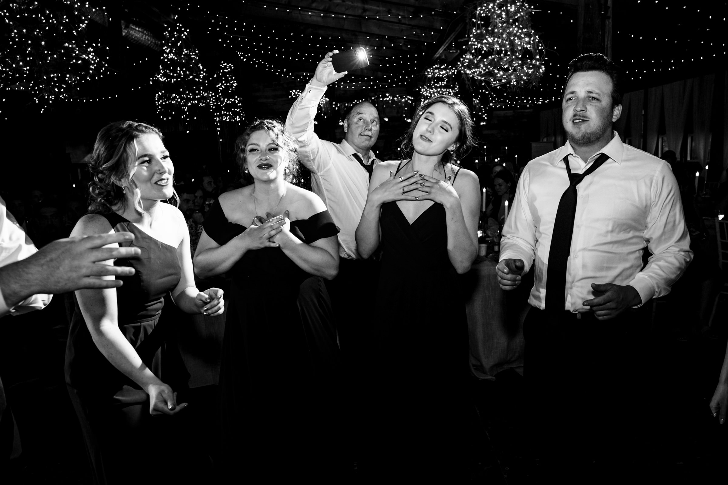 People in formal wear sing and dance at a Winnipeg wedding venue under twinkling lights.
