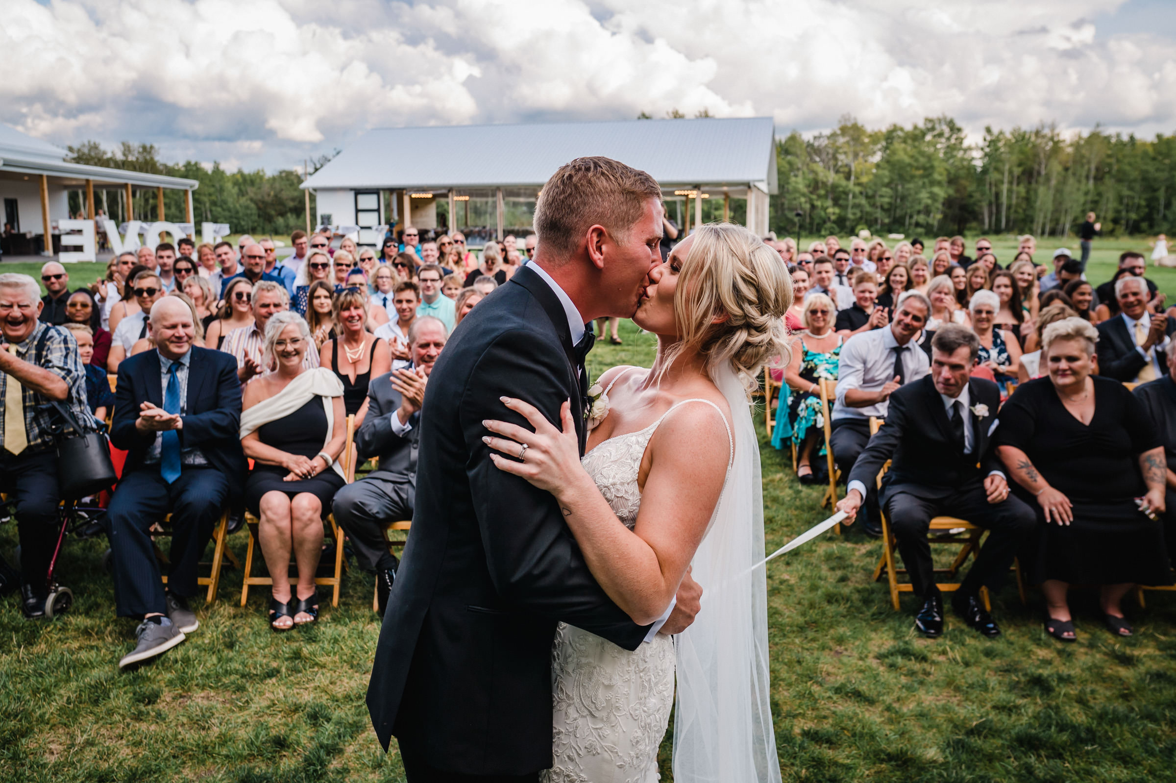 Bride and groom kiss at a Winnipeg wedding venue before a seated outdoor crowd.
