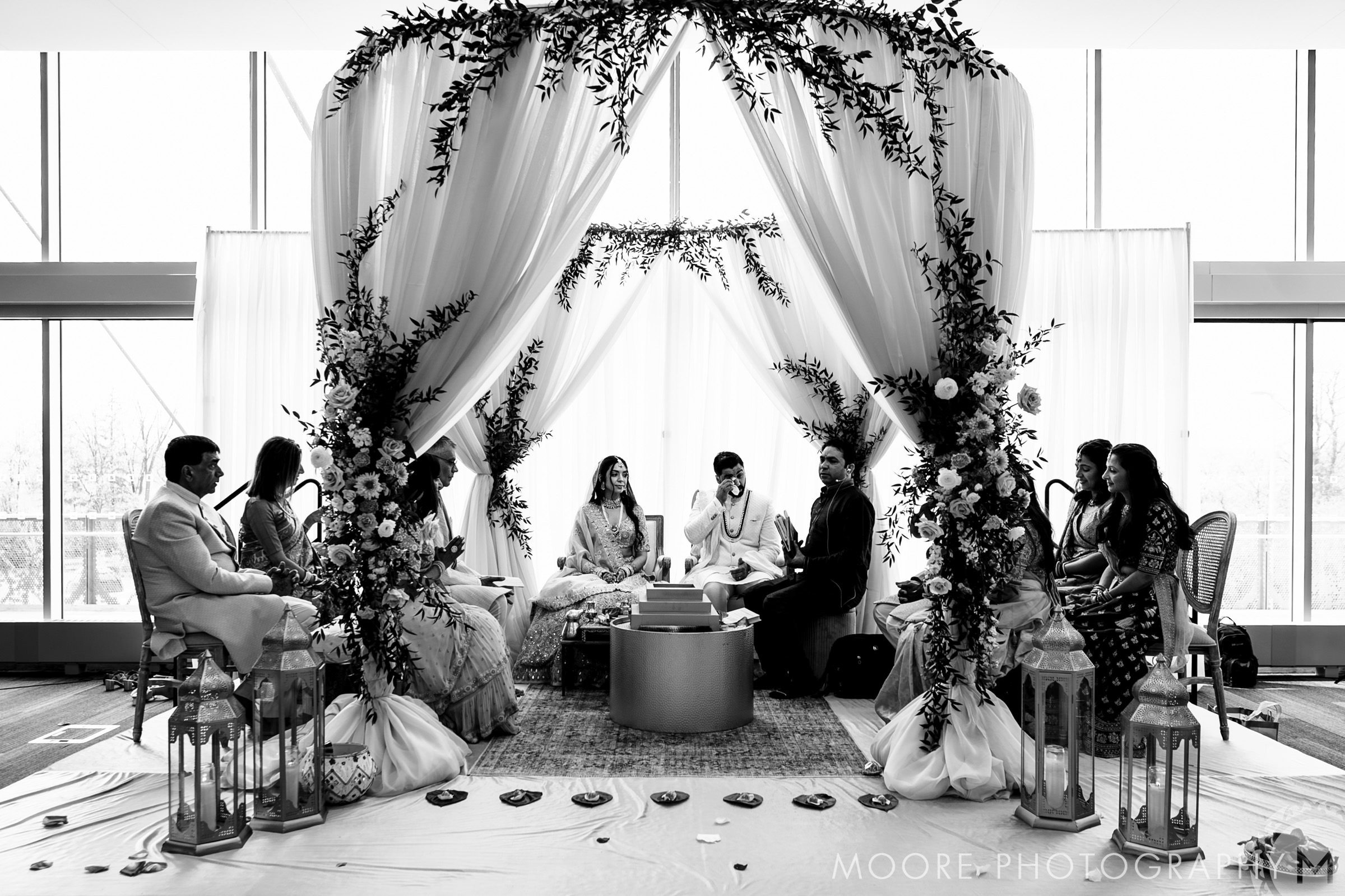 A wedding couple is seated beneath a floral canopy, surrounded by family in Winnipeg venues.