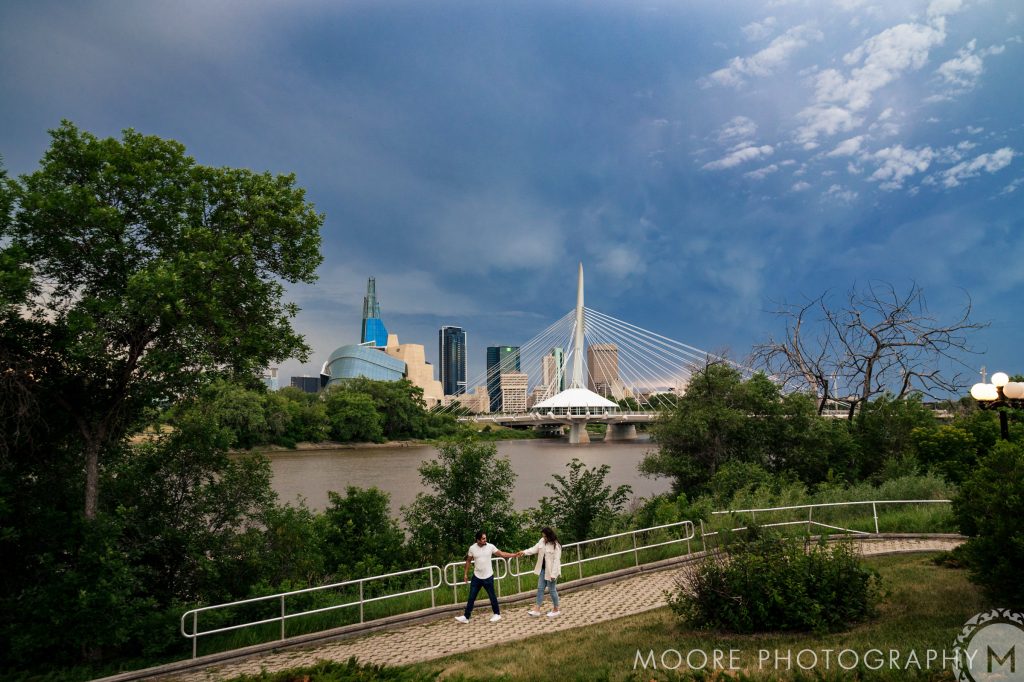 Engaged couple, holding hands and walking down a path with the Winnipeg skyline in the background