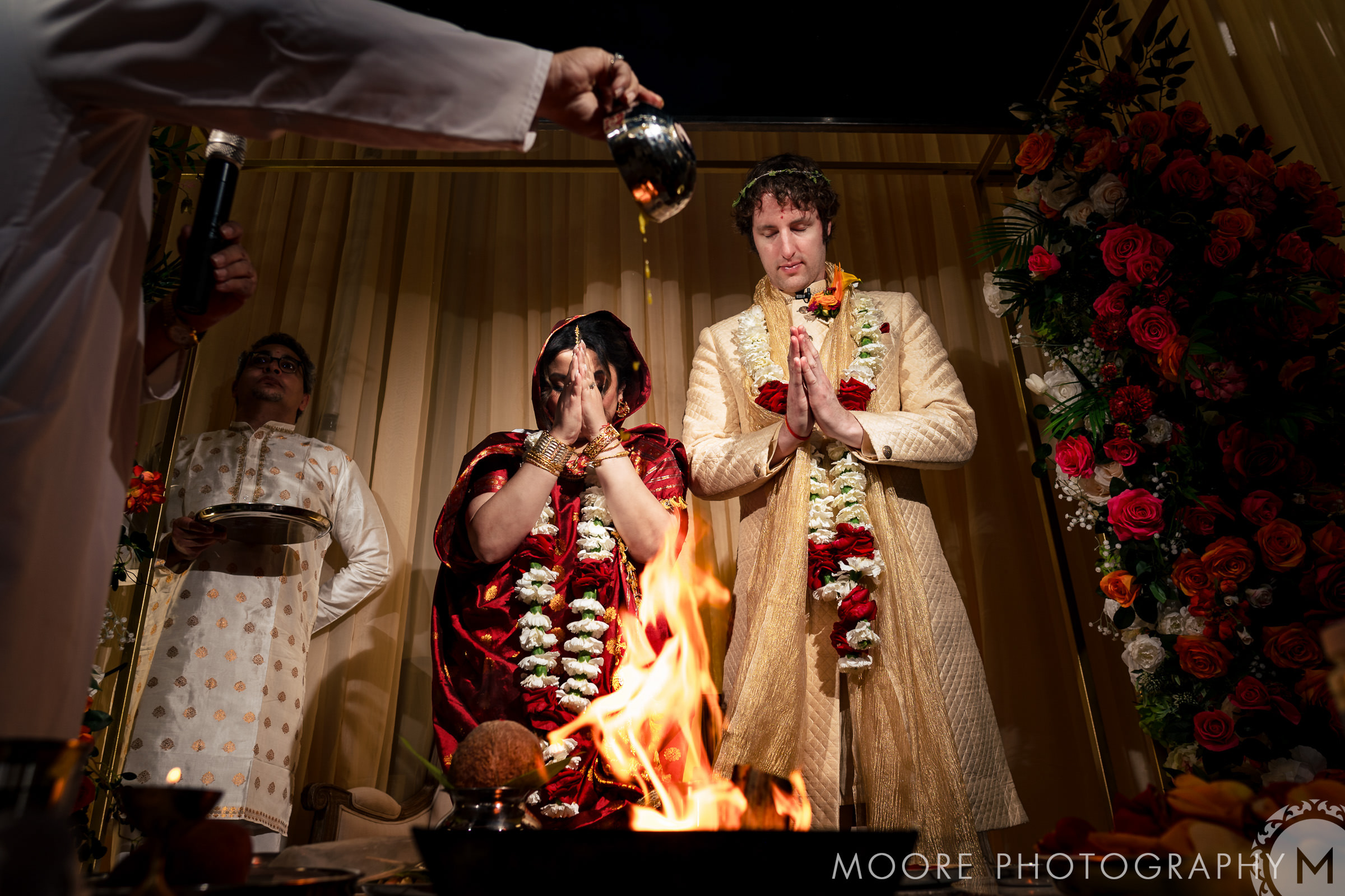 A bride and groom in traditional attire participate in a wedding ceremony. The bride, wearing a red saree, and the groom, in a cream sherwani, both have floral garlands. A person pours a liquid into a ceremonial fire. Floral decorations surround the scene.