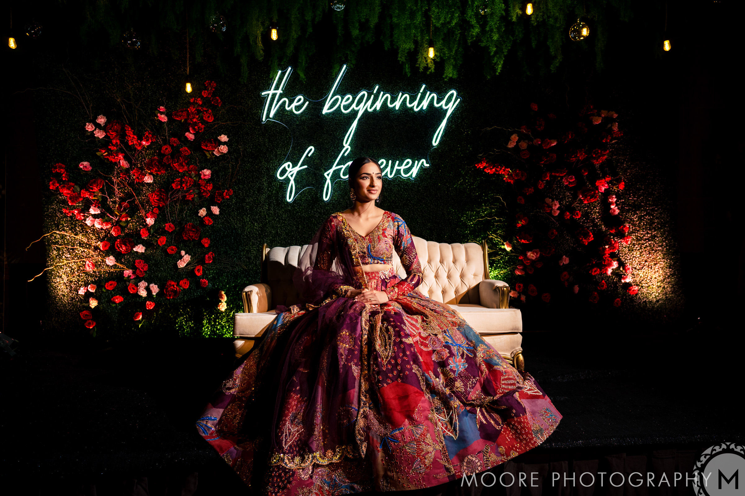 Indian bride in a colorful gown seated on a sofa with neon sign and floral decorations.