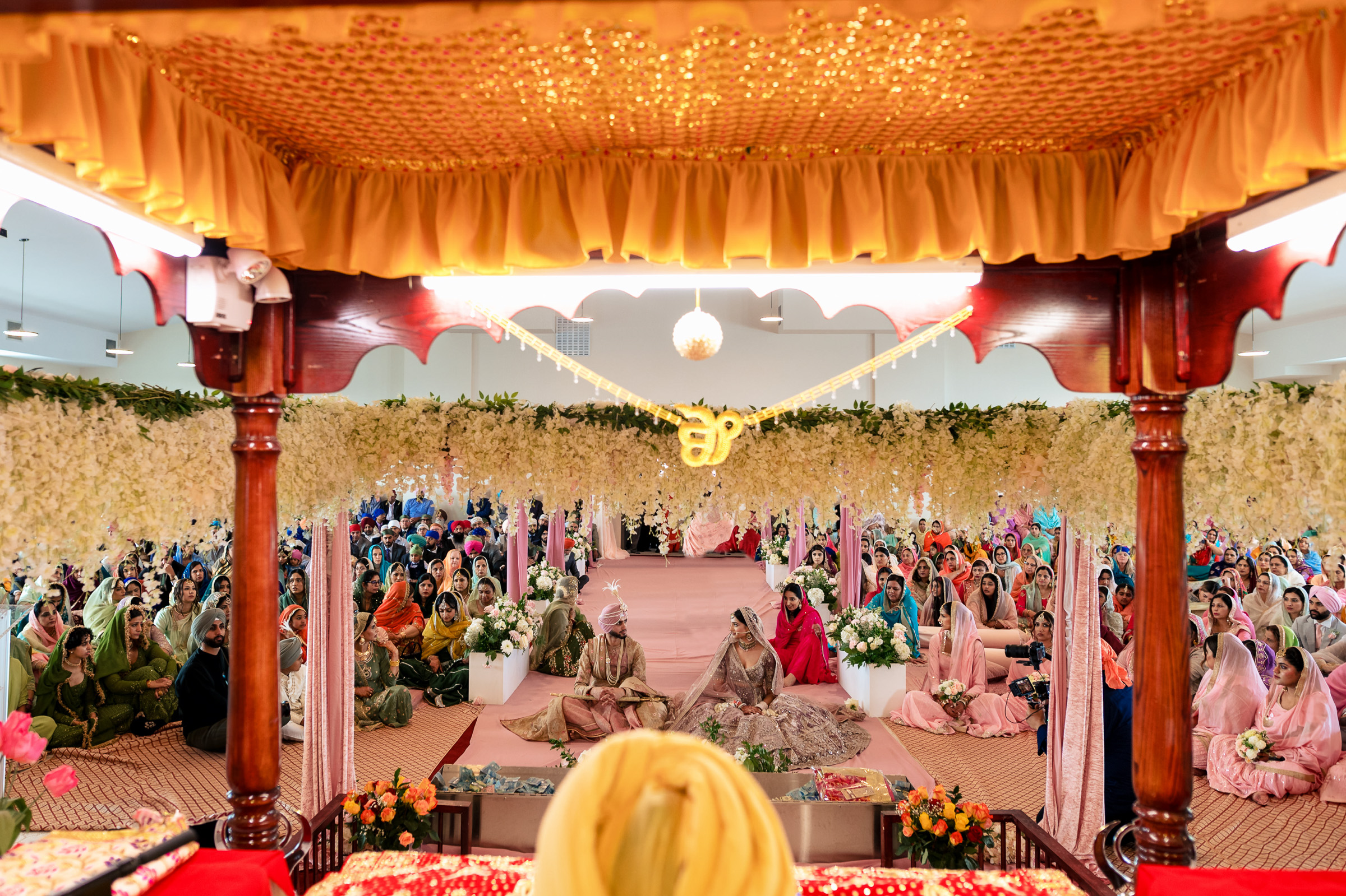 People gathered in a decorated hall for a Sikh wedding, seated on the floor in colorful attire.