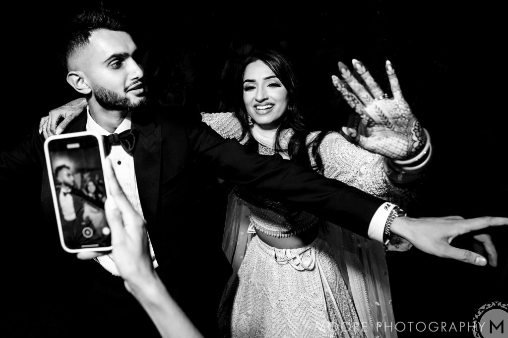 A couple dancing at a Winnipeg wedding venue, smiling; woman with hennaed hands; phone capturing moment.
