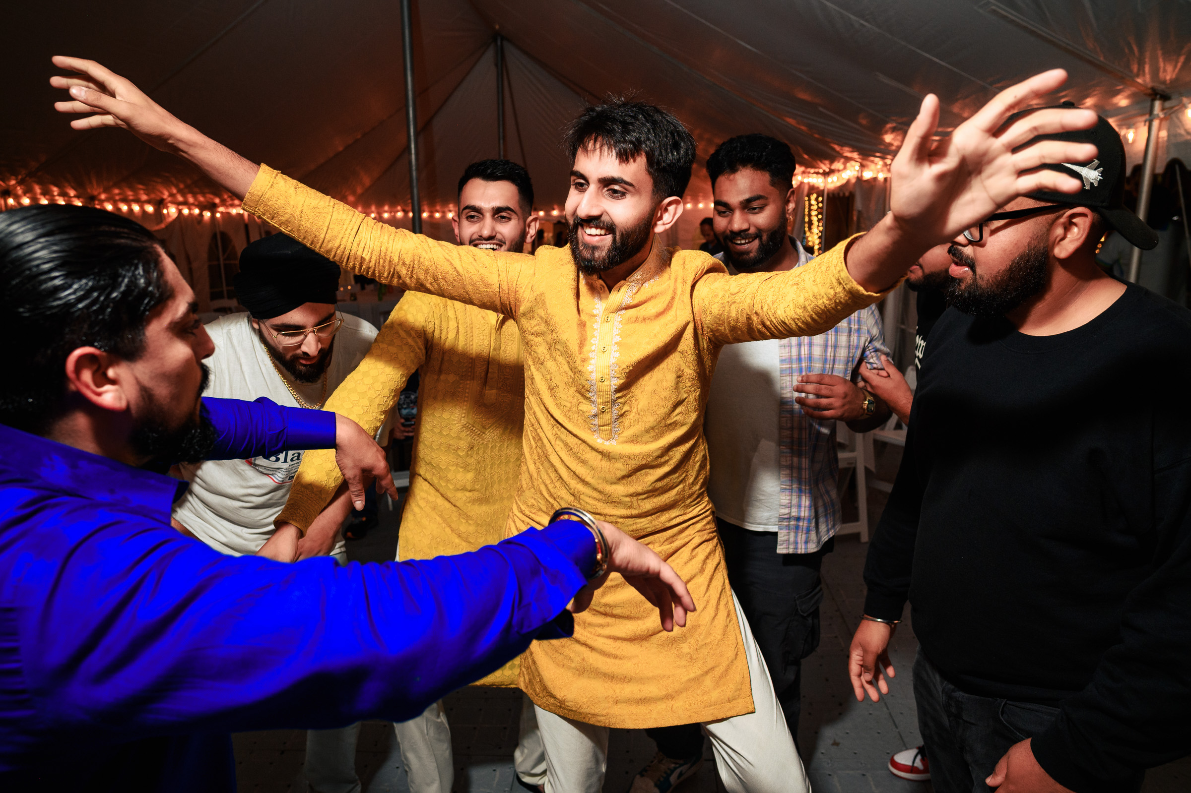 Man in yellow kurta dancing joyfully with friends at an Indian wedding tent.