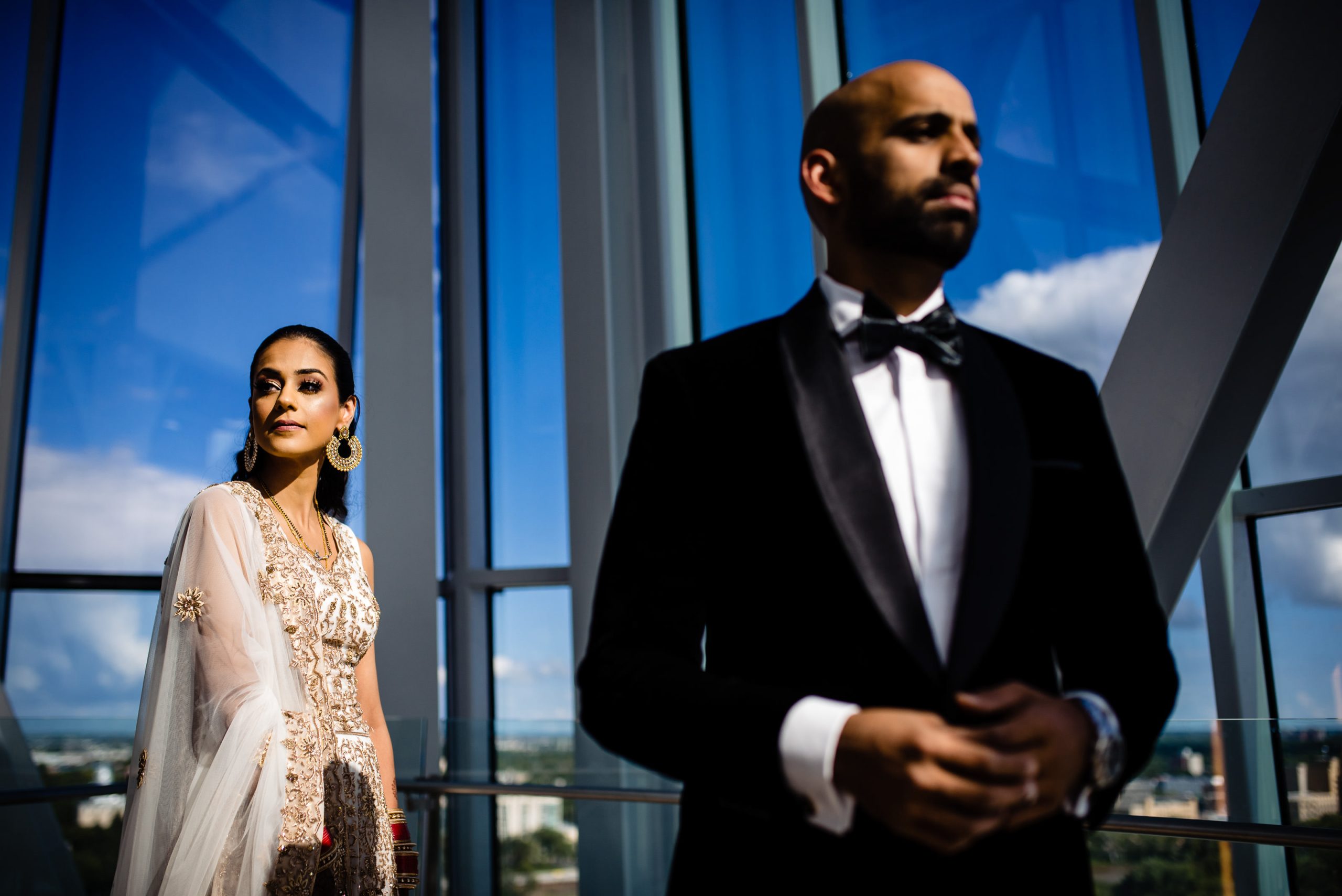 A woman in traditional attire with gold embroidery stands near a window, gazing to the left. A man in a black tuxedo and bow tie stands in the foreground, facing right. The background shows a bright sky through tall glass windows.